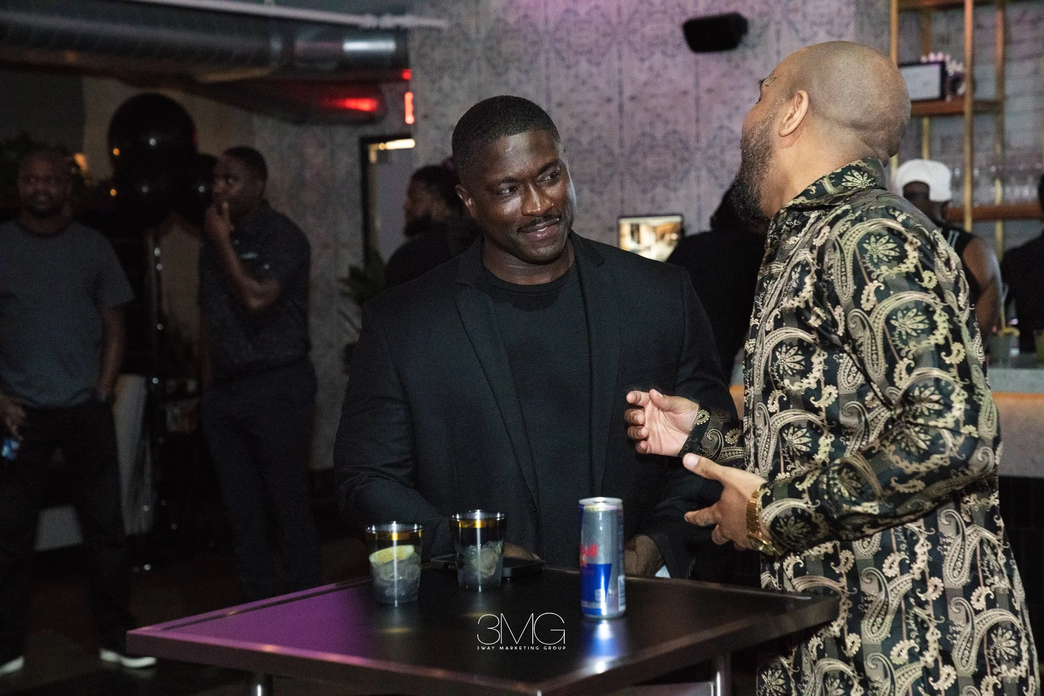 Two men are engaged in a handshake or hug at a social event in a dimly lit room. One man is dressed in a dark blazer and shirt, while the other is wearing a patterned shirt. There are several glasses with drinks on the table in front of them, and peo