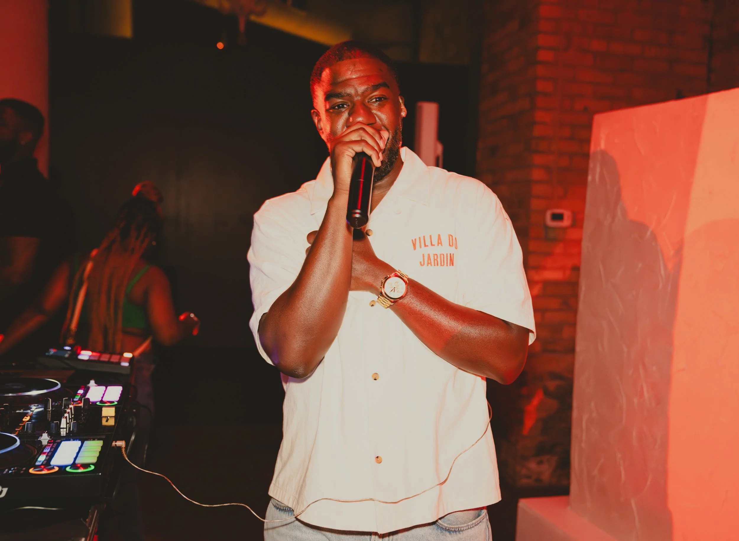 A male DJ standing in a dimly lit venue, holding a microphone and wearing a white shirt with words 'Villa Di Jardin' on it. There is DJ equipment with colorful lights in front of him and a brick wall in the background.