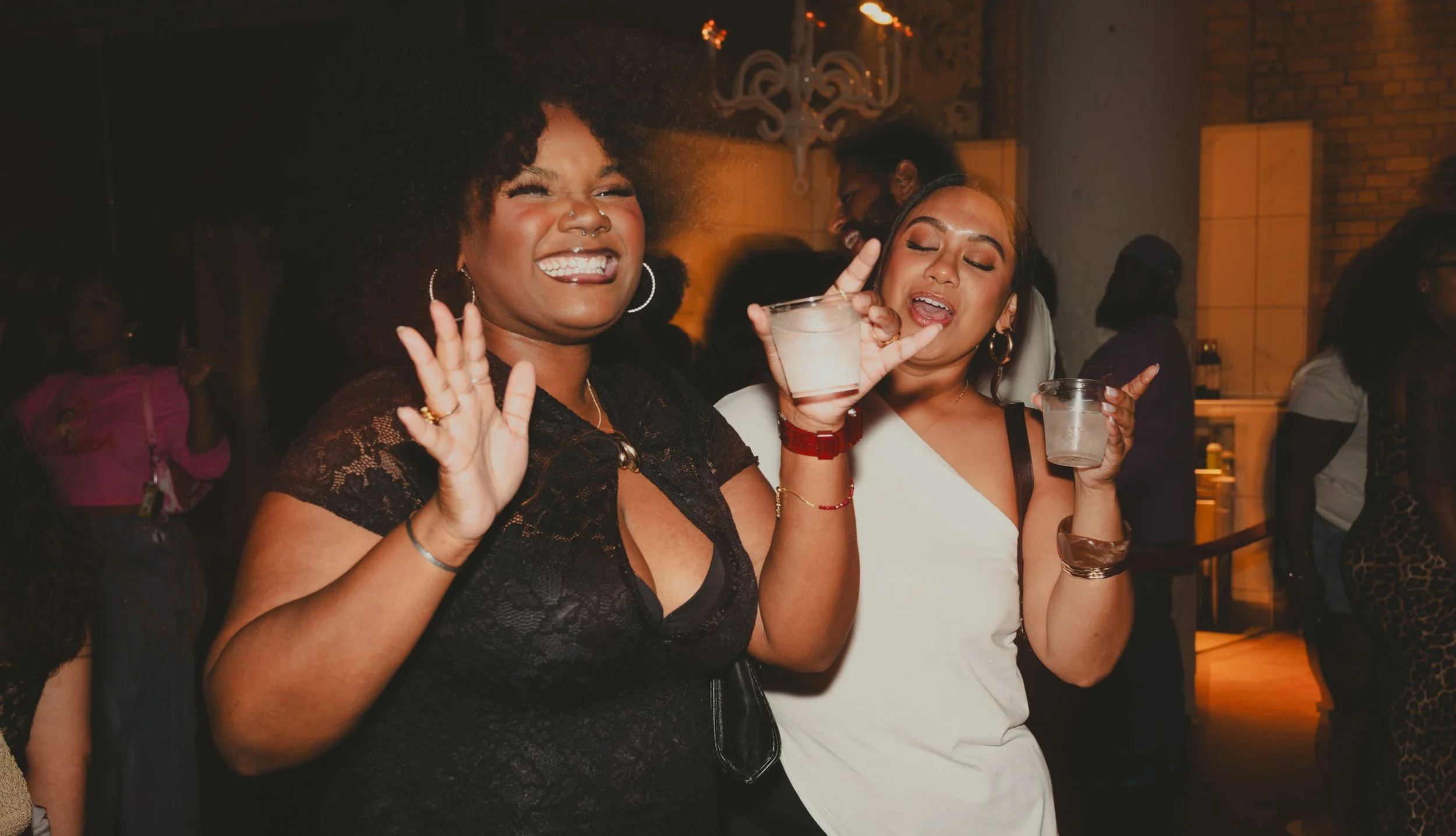 Two women dancing and enjoying drinks at a lively indoor party with other people in the background.