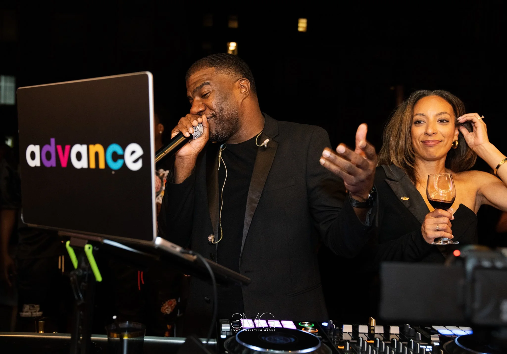 A man and woman at a DJ booth at a nighttime event. The man is singing into a microphone, and the woman is smiling with a glass of wine in her hand. The DJ setup includes a laptop with "advance" logo and DJ equipment.