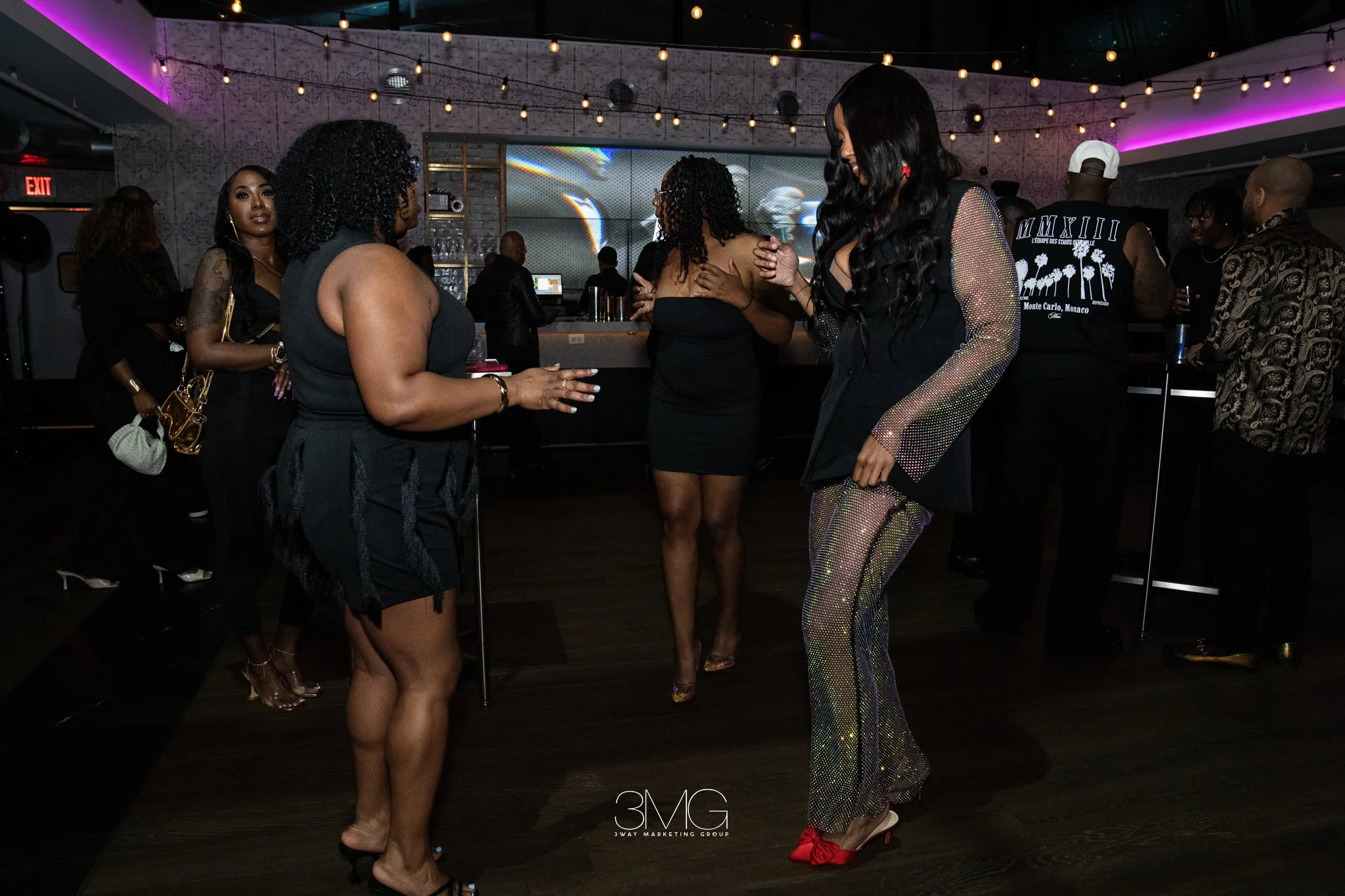People dressed in fashionable outfits standing inside a nightclub or lounge with purple and pink lighting, some engaged in conversation or dancing