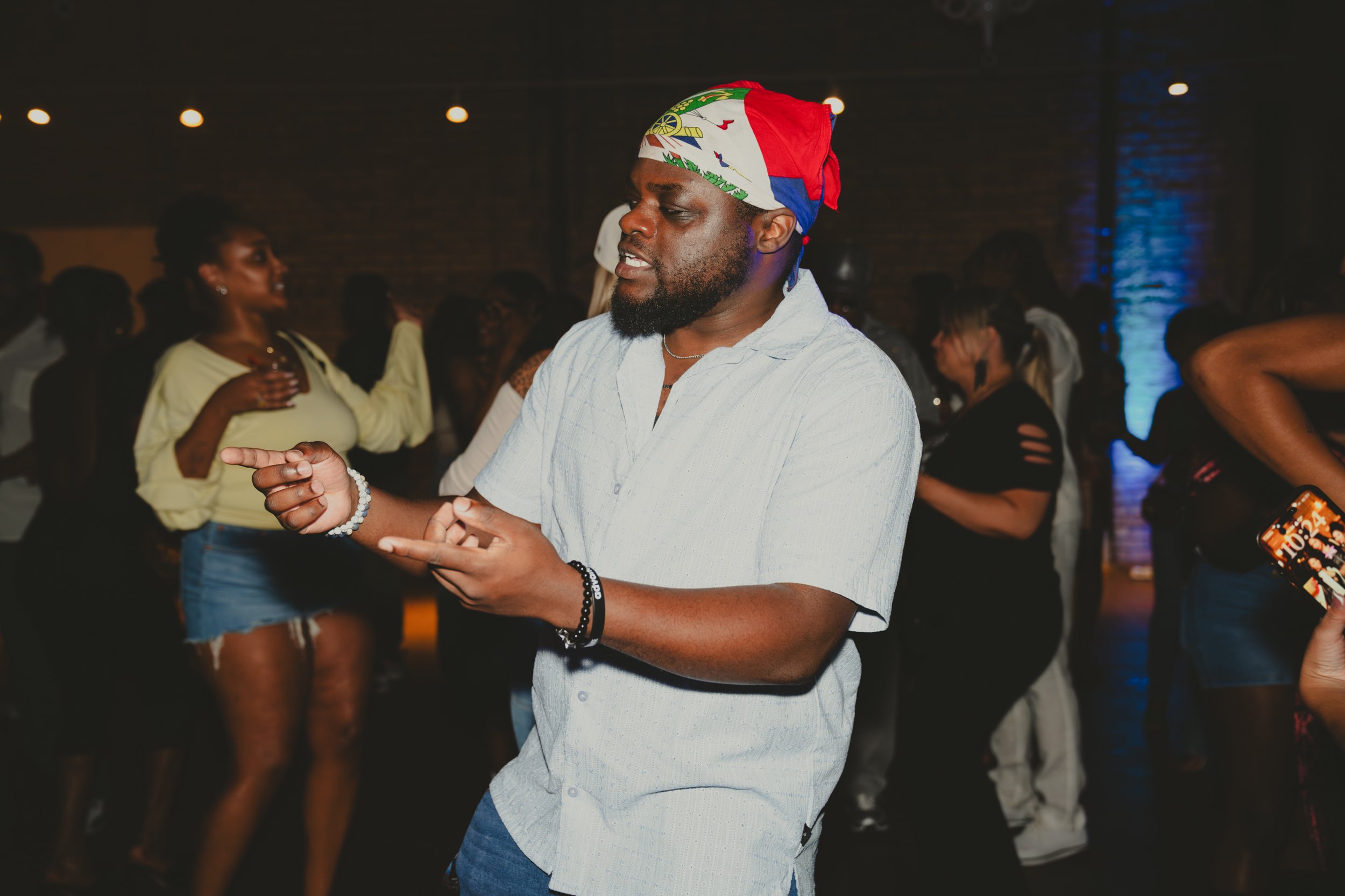A man dancing at a party wearing a colorful bandana and a light-colored shirt. In the background, people are socializing and enjoying the event in a dimly lit, urban-style venue.