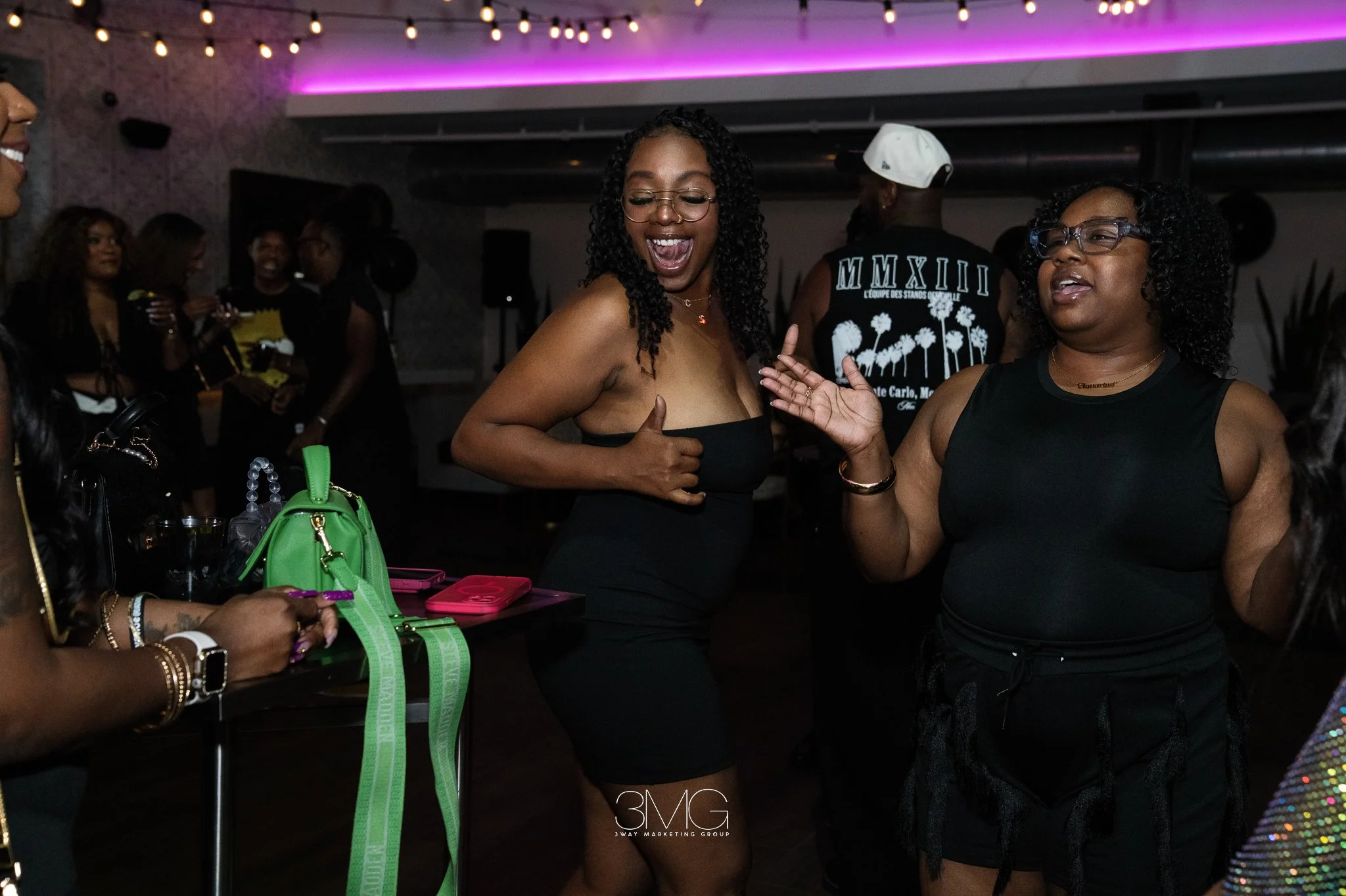 Group of people dancing and socializing at a party or nightclub with colorful lighting and string lights overhead.