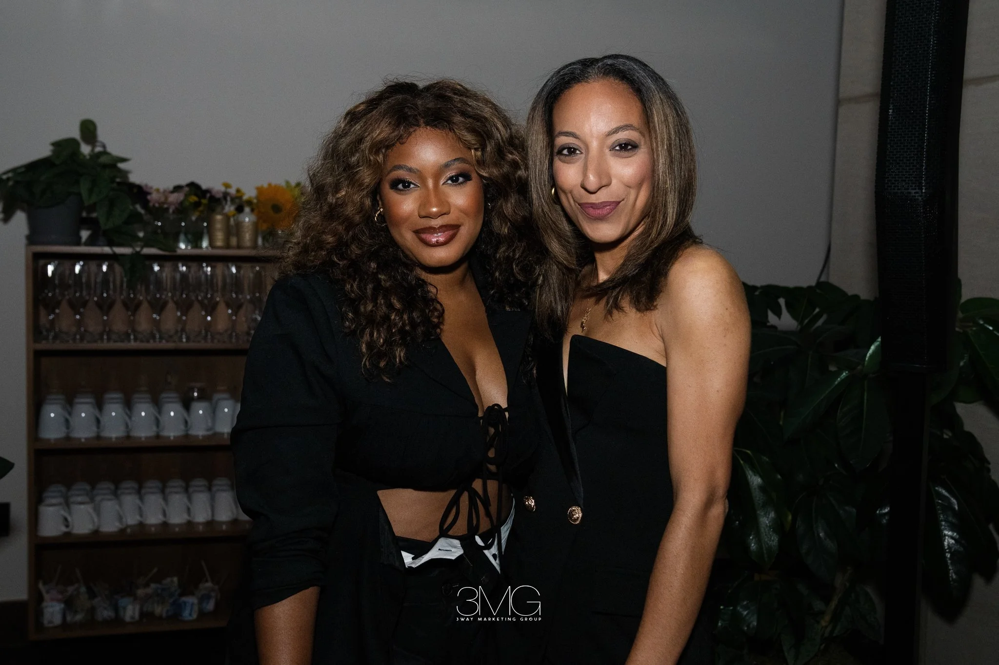 Two women standing closely together at an event, smiling for the camera, with a backdrop of plants and glasses on shelves.