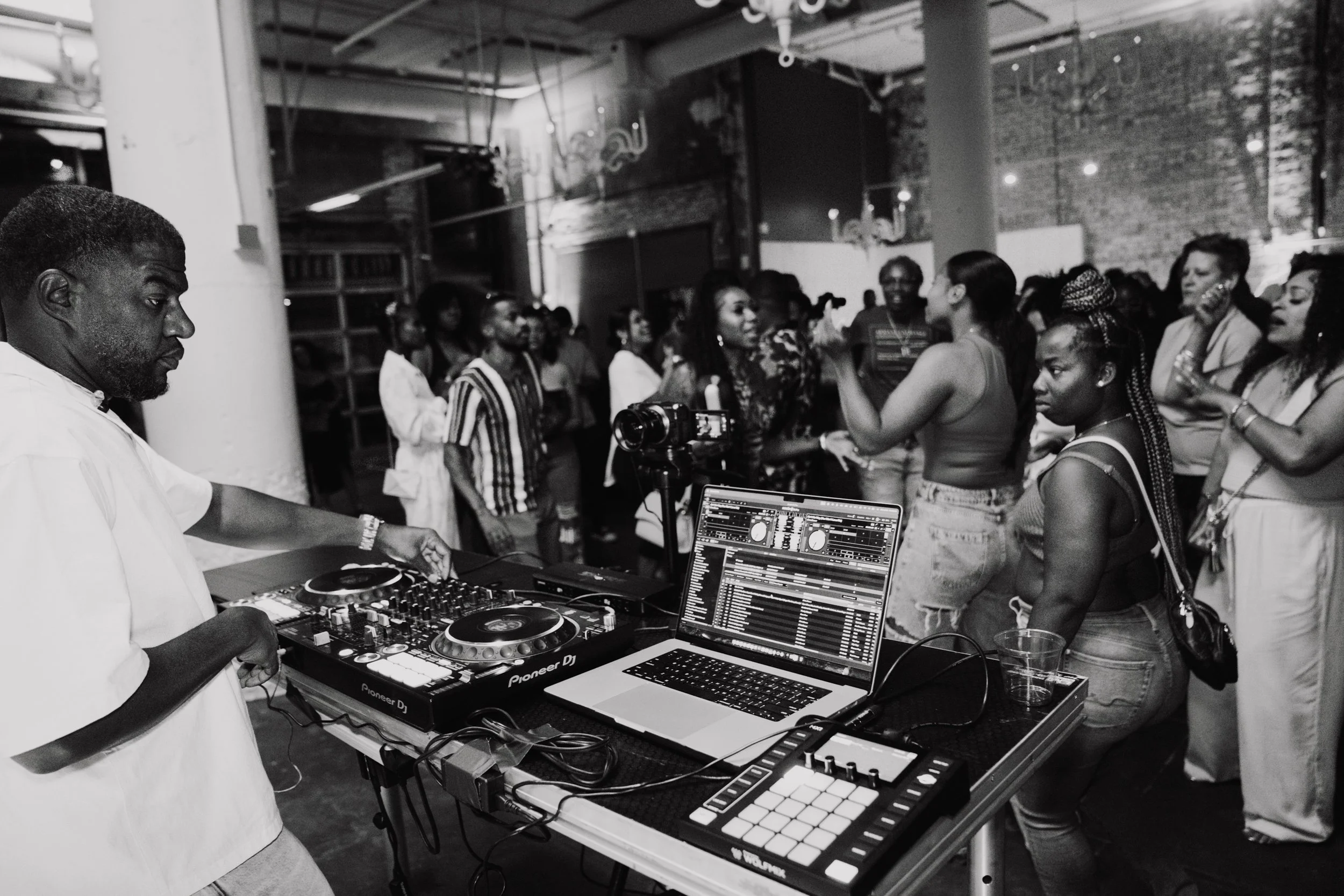 A DJ is mixing music on a Pioneer DJ controller, with a laptop displaying a DJ software interface in front of him. He is in a crowded indoor venue with exposed brick walls, and people are socializing and dancing, enjoying the music.