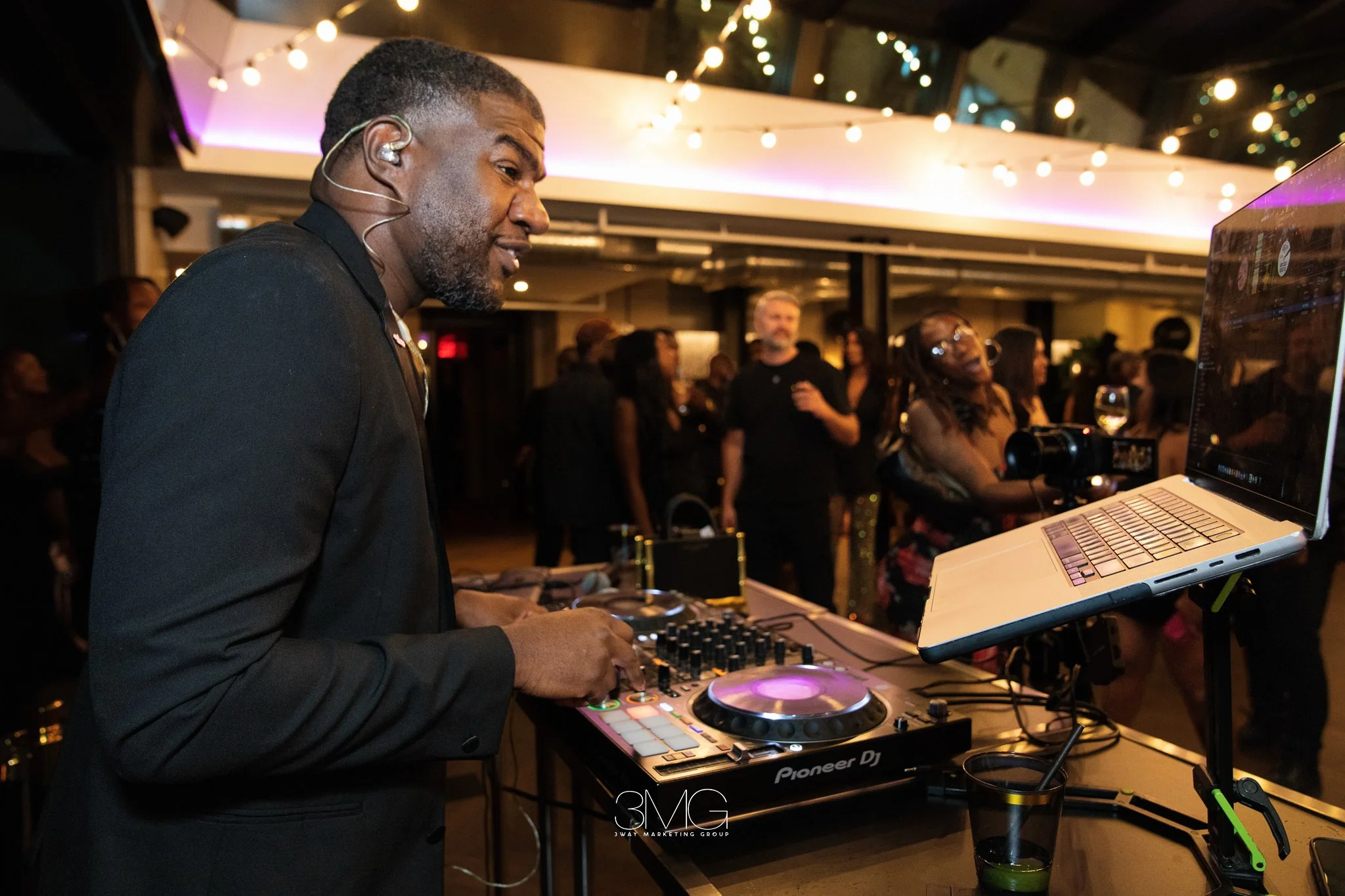 A DJ is performing at a party, using a Pioneer DJ controller and a laptop, with a crowd of people dancing and socializing in a dimly lit venue decorated with string lights.