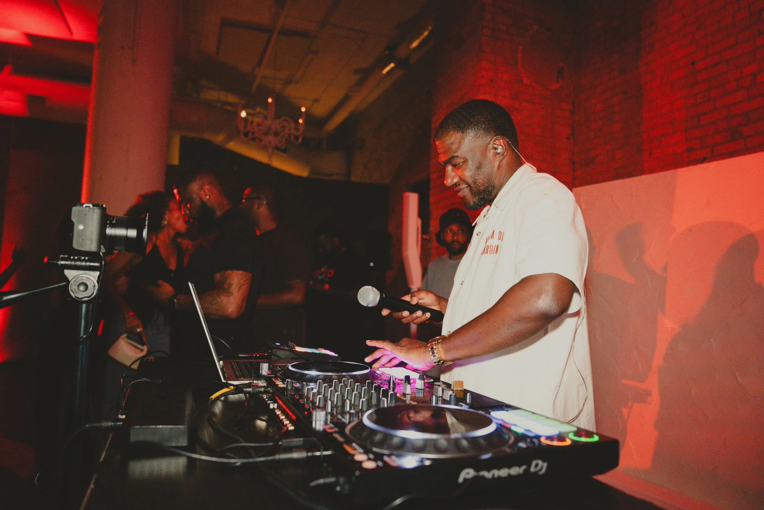 A DJ is performing at a party or event, standing behind a Pioneer DJ setup with a mixer and turntables, holding a smartphone. There are people in the background with dim lighting and red hues, and a chandelier is visible on the ceiling.