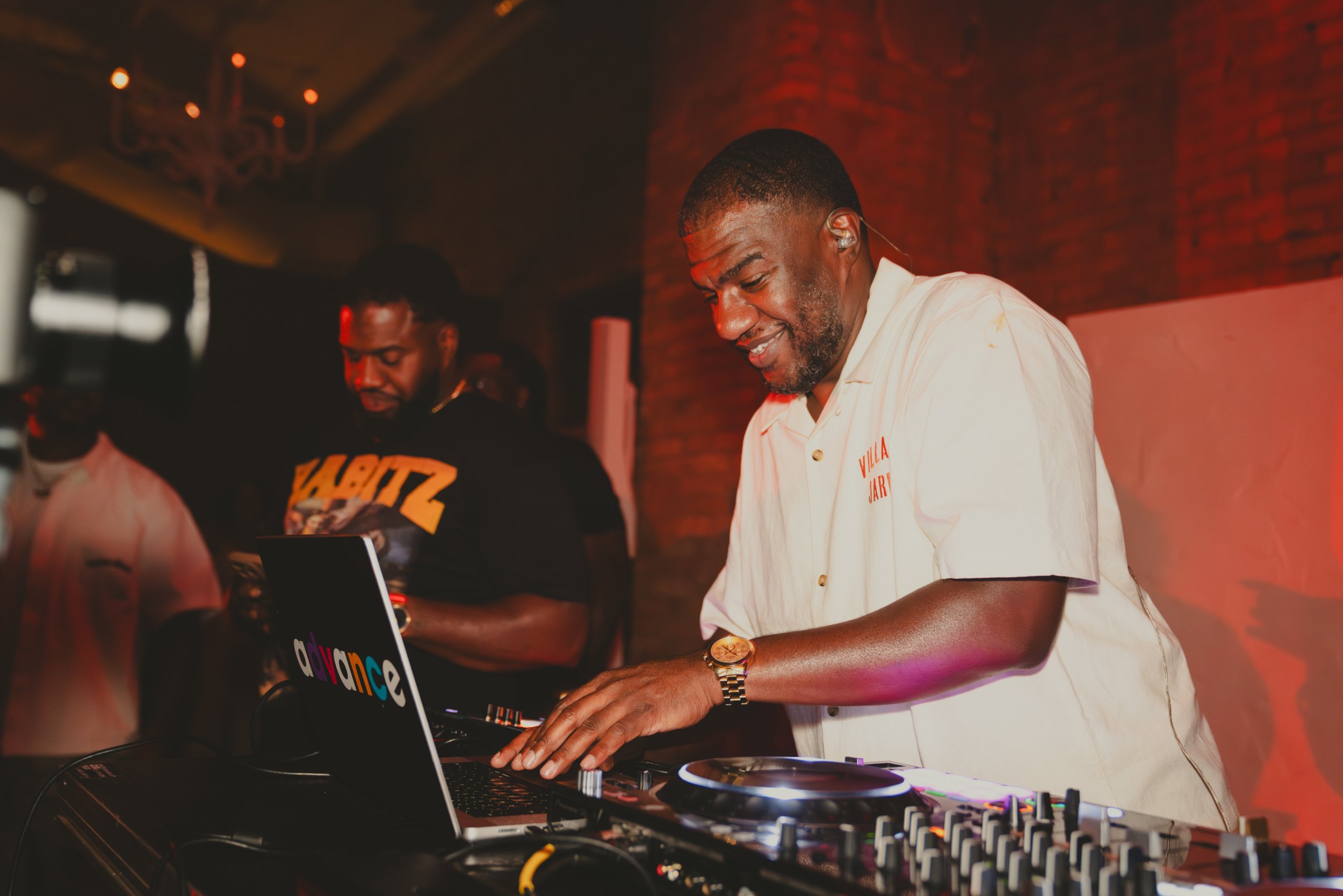 Two men are DJing at a lively event in a dimly lit room with a red brick wall. The man in the foreground is smiling, wearing a cream-colored shirt with orange text, a gold watch, and a headset. The second man in the background is wearing a dark hoodi