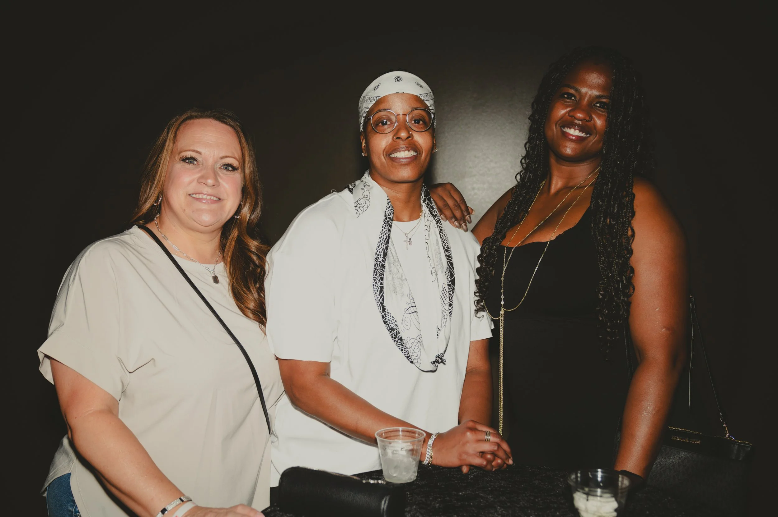 Three women standing together at an event, smiling, with drinks on a table in front of them, against a dark background.