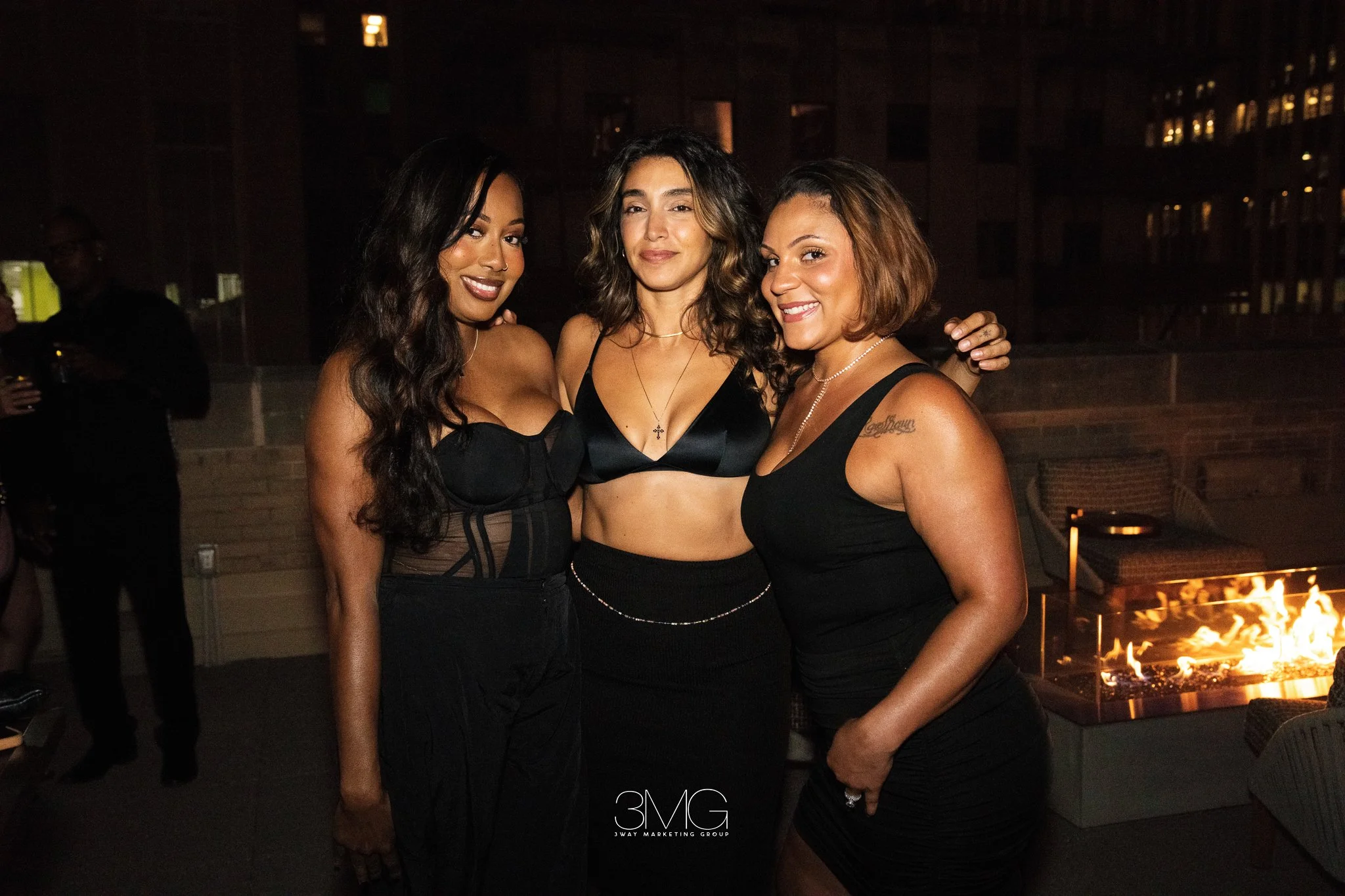 Three women posing together at an outdoor nighttime gathering with a fire pit in the background.