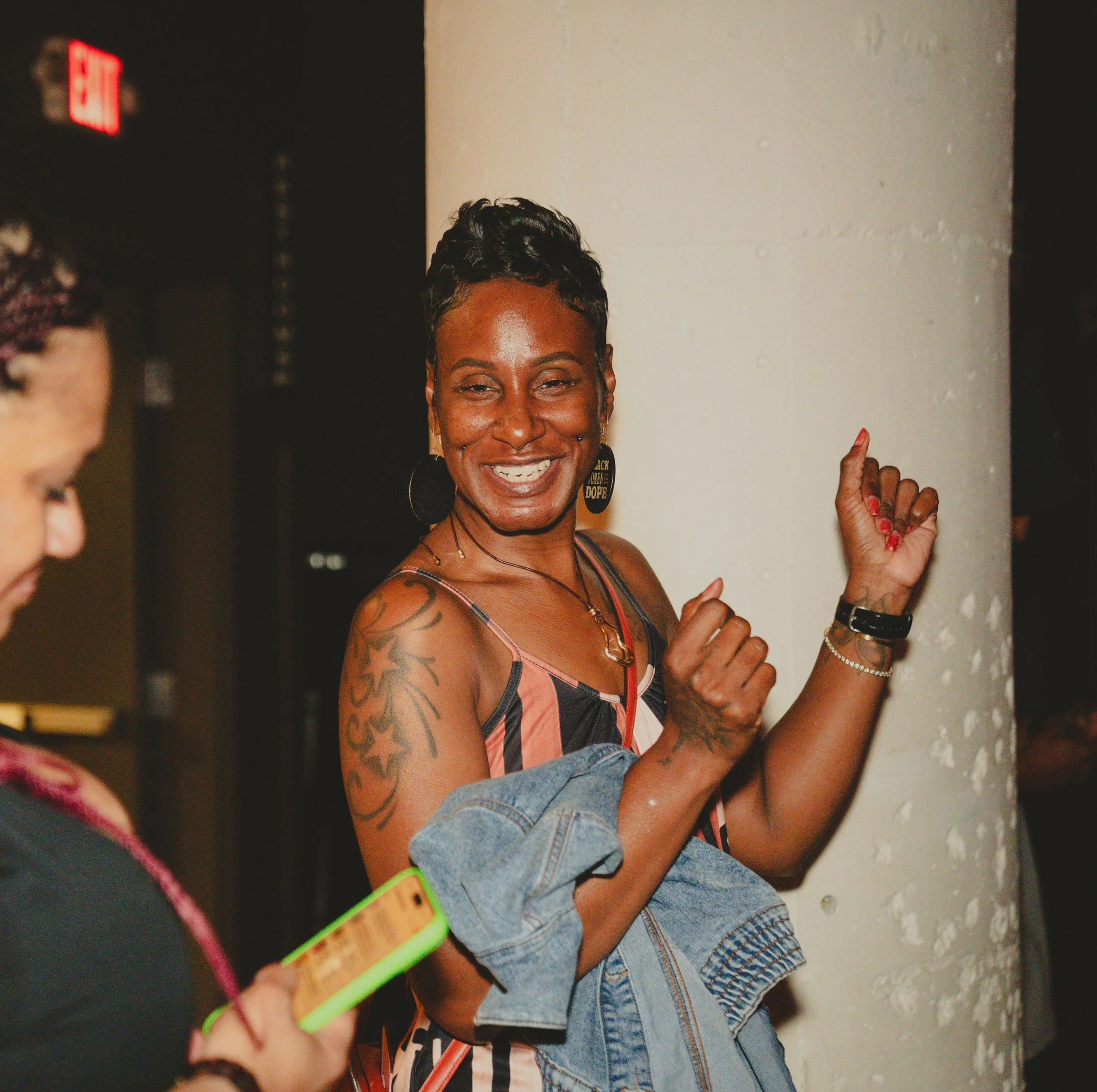 A woman with short black hair styled in a mohawk, smiling, raising her right fist, tattoos on her right arm, wearing earrings and a multicolored top, standing next to a white wall.