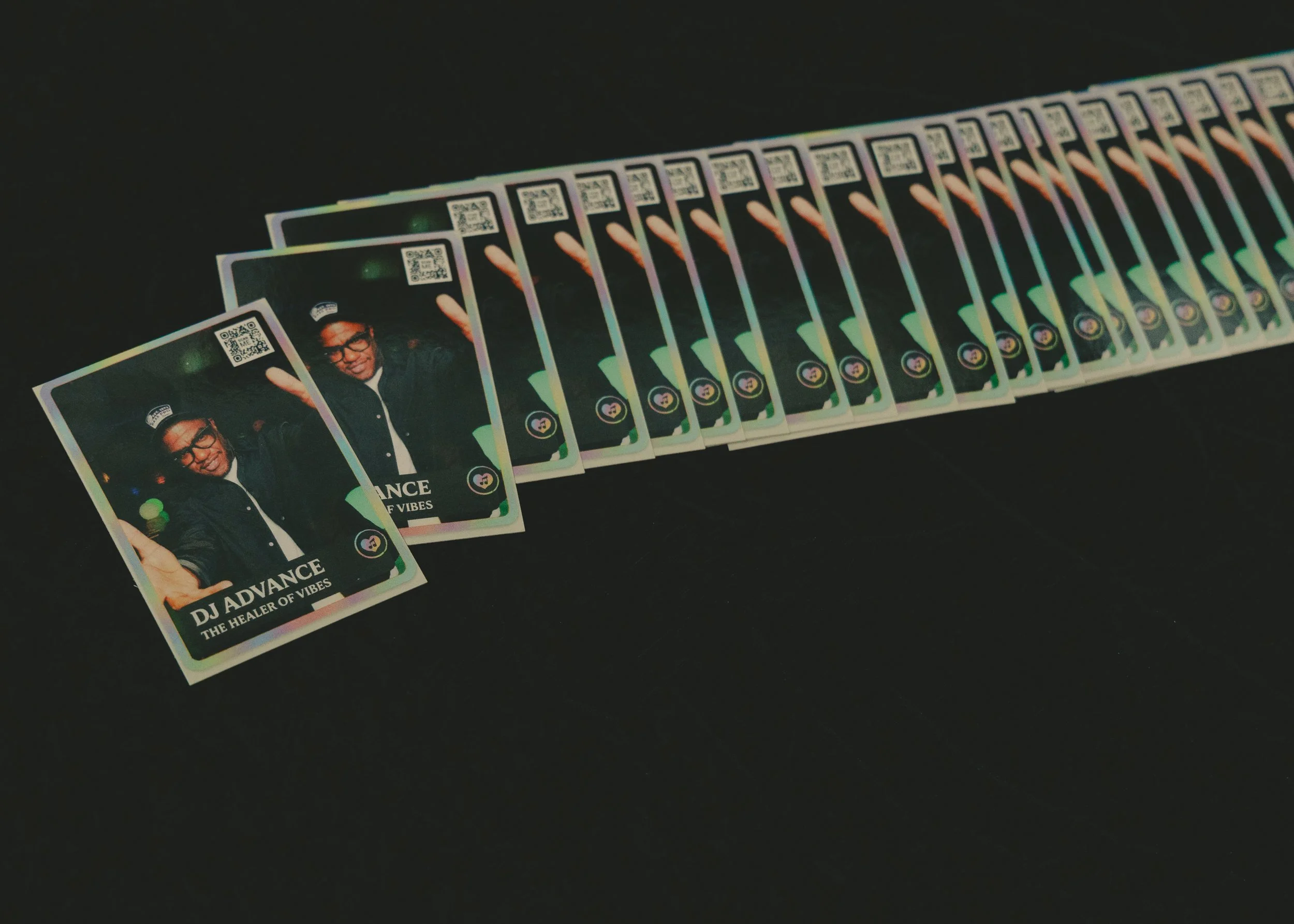 Several holographic business cards spread out on a dark surface. The cards feature a photo of a man wearing glasses and a cap, with text that reads 'DJ ADVANCE' and 'THE HEALER OF VIBES'.