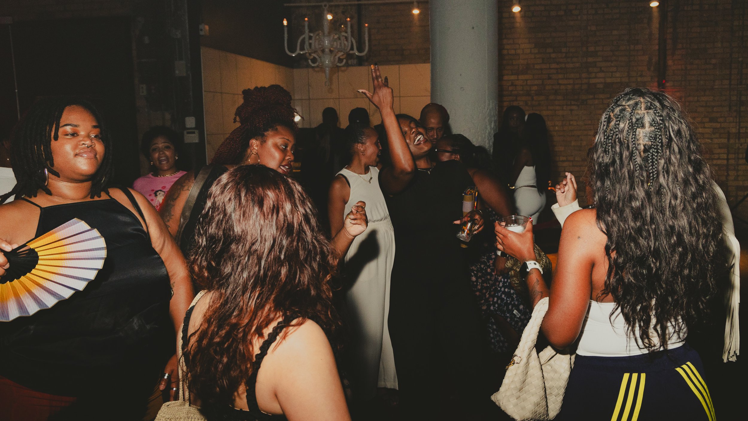 A group of women at a party or event, some wearing dresses, with one woman dancing and others holding drinks, in a dimly lit room with brick walls.