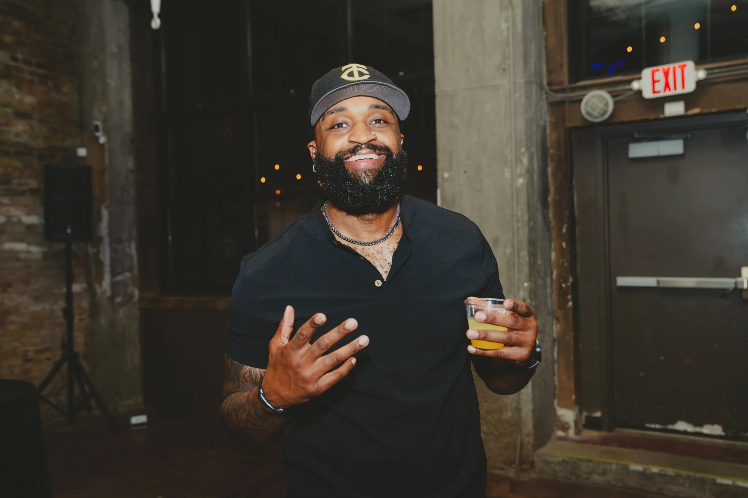 A man with a beard, nose piercing, and tattoos, holding a drink, smiling at the camera in an indoor space with brick walls and an exit sign in the background.
