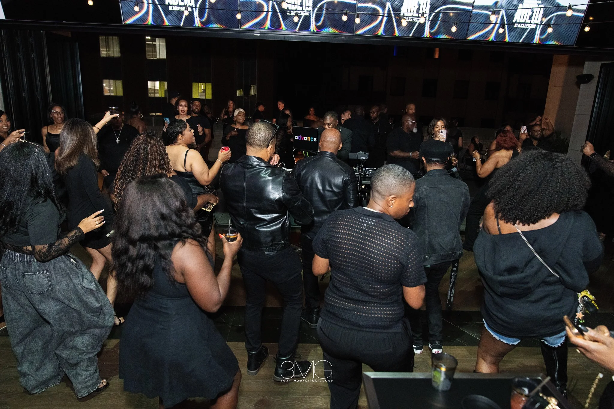 Crowd of people gathered at a social event or party in a dimly lit indoor space with some individuals holding drinks and taking photos.