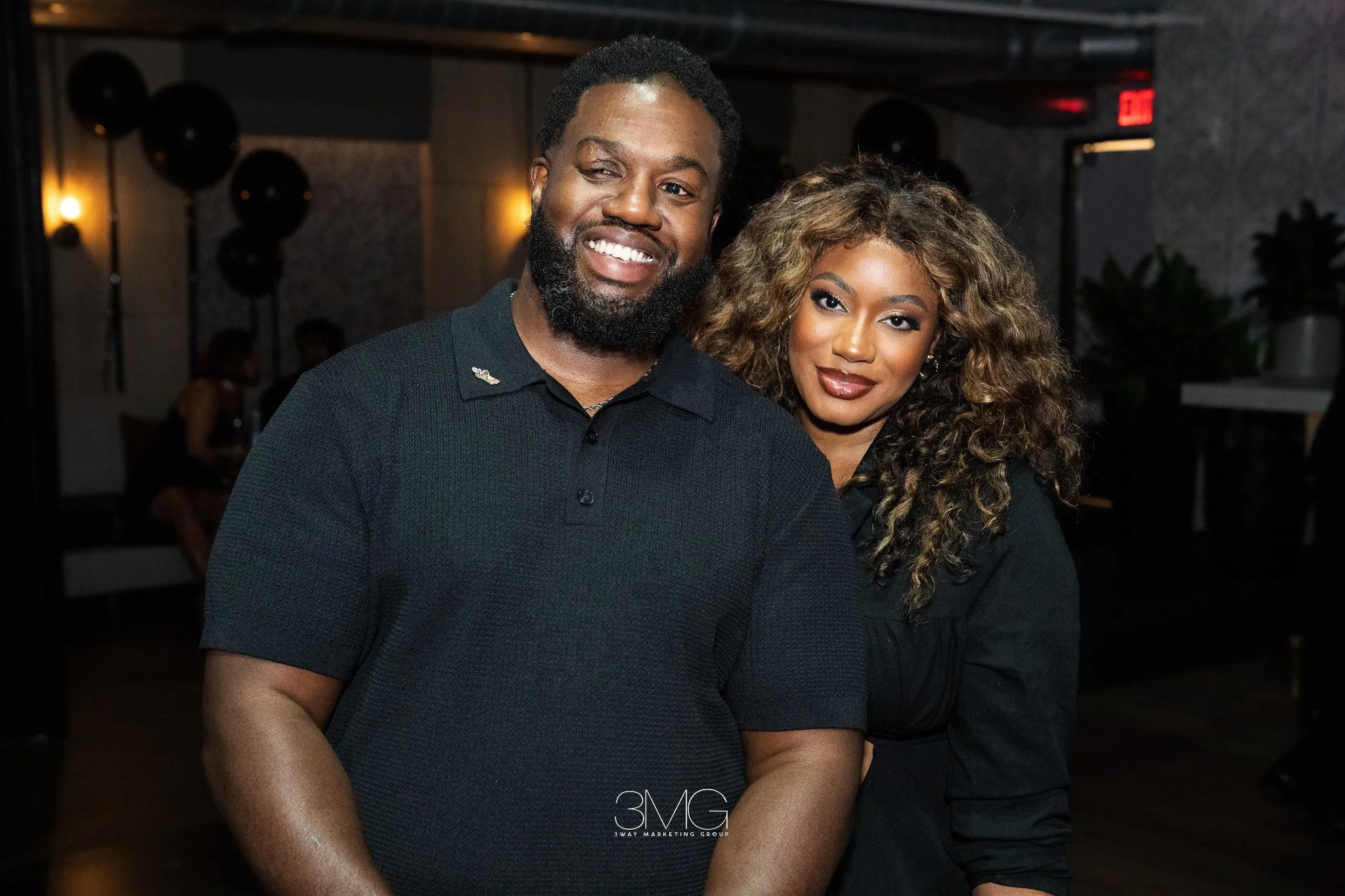A smiling Black man and woman posing together at an indoor event or party, with dark background and ambient lighting.
