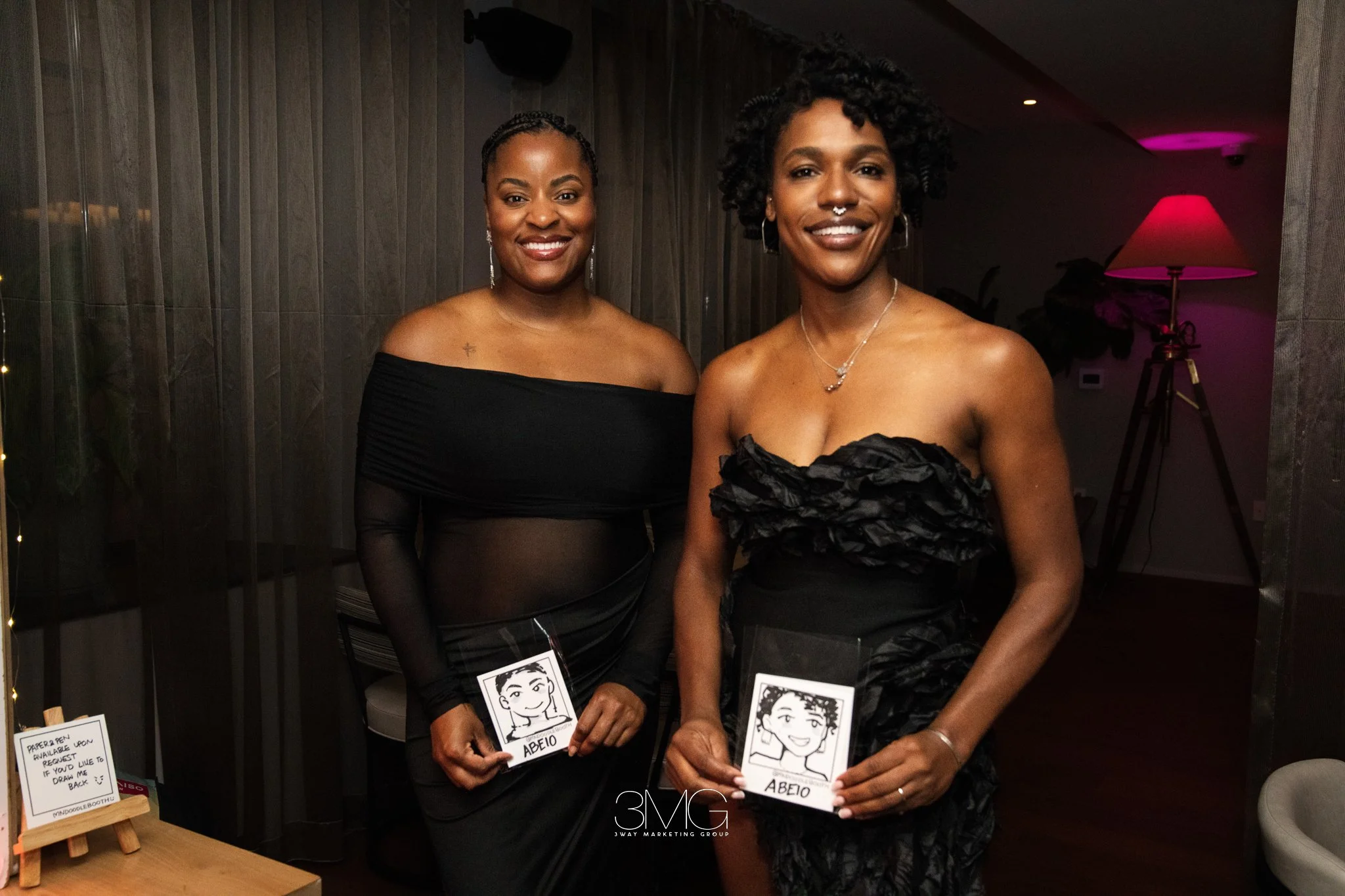 Two women dressed in black at an indoor event, smiling and holding illustrated signs with the name "ABEO." One woman has short curly hair and a septum piercing, and the other has braided hair. The background includes a standing lamp with a pink shade