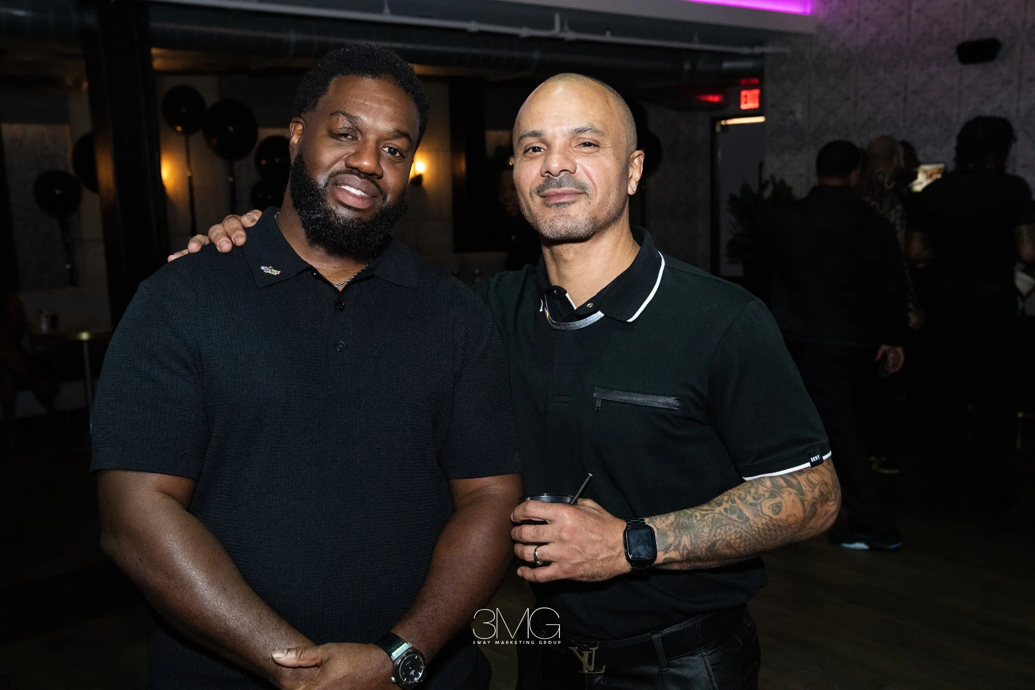 Two men standing close together at a social event, one with a dark complexion and beard, the other with a light complexion and tattoos on his arm, both smiling slightly.