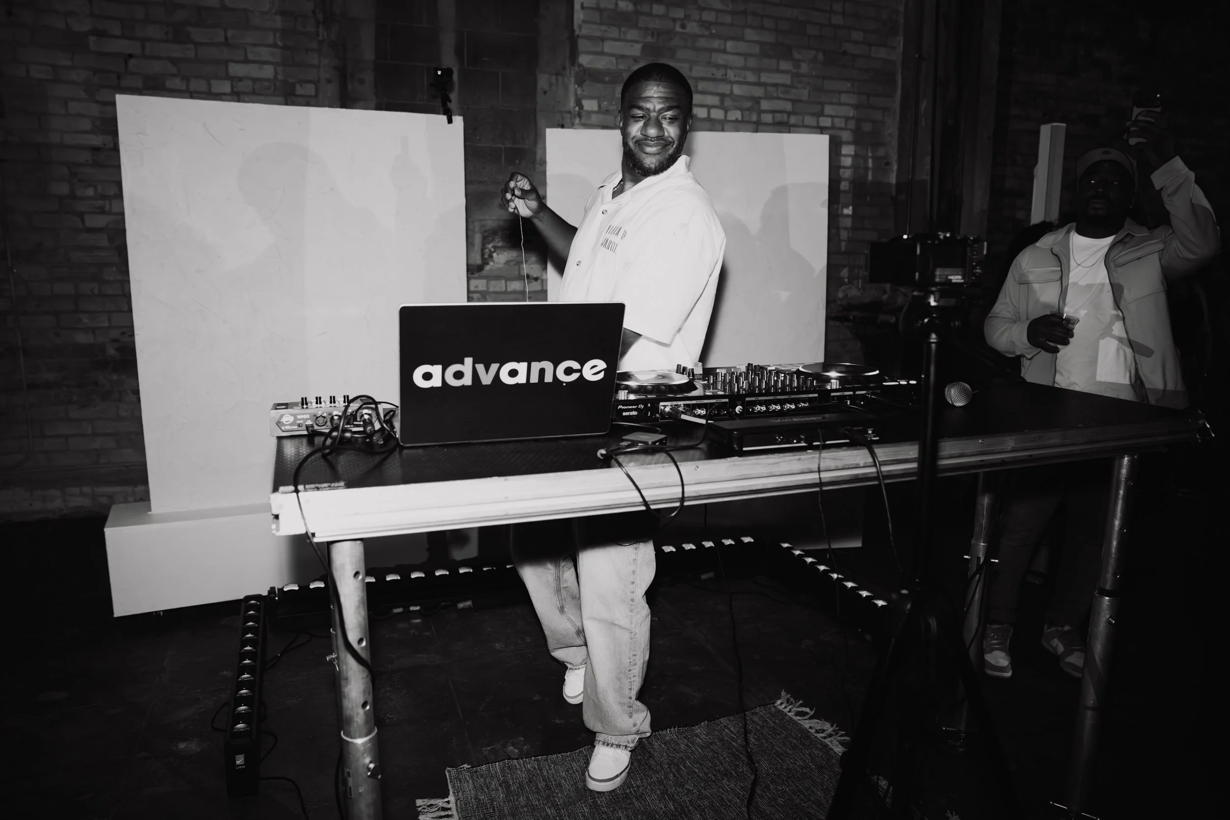 A man with a beard and a smile, wearing a white shirt, is DJing at a party with a laptop that has the word 'advance' on it. He is standing behind a DJ console, holding a headphone in one hand. There is another person in the background wearing a light