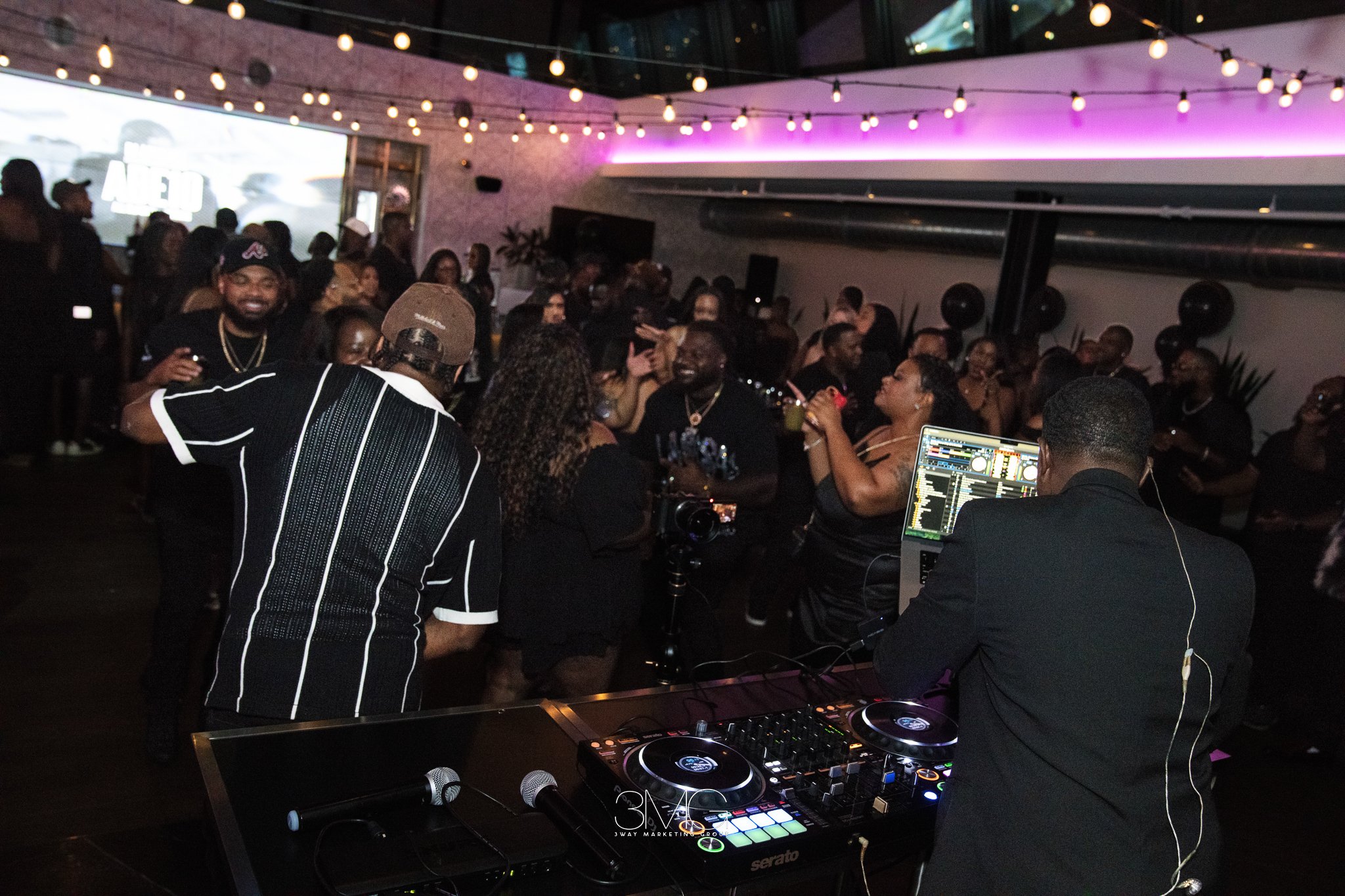 A lively indoor party scene with people dancing and socializing. A DJ stands at a mixing console with turntables, wearing headphones, in the foreground. The room is decorated with string lights and purple ambient lighting, creating a festive atmosphe