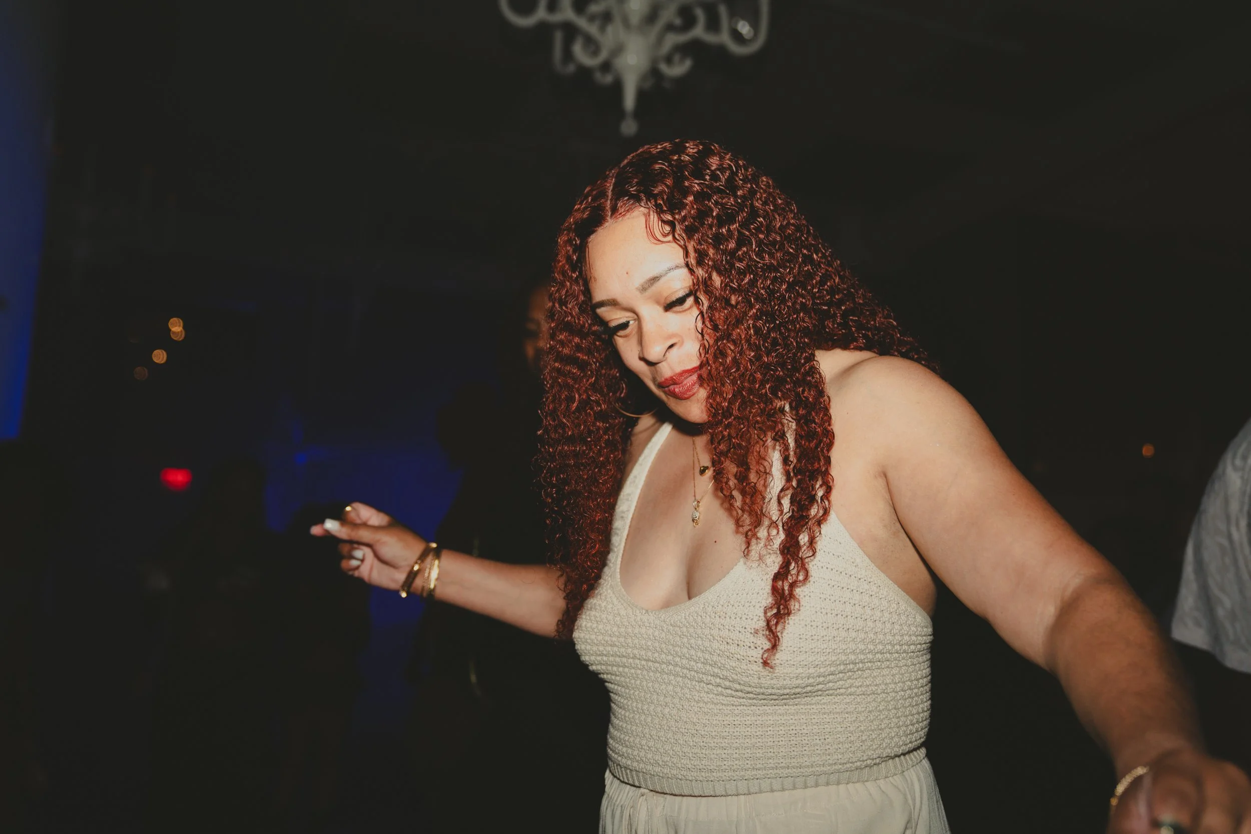 A woman with curly red hair wearing a beige sleeveless top and white skirt, dancing with arms raised in a dark setting.