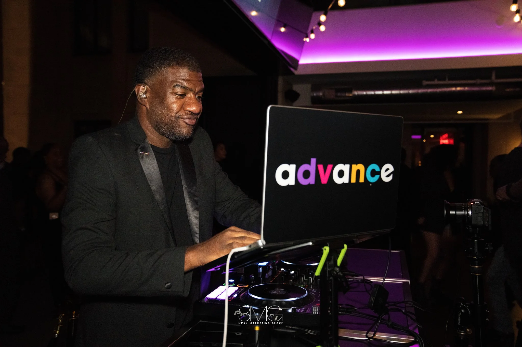 A DJ with short hair and a beard, wearing a black jacket, is performing at an event. He is using a laptop with a colorful 'advance' sticker and DJ equipment, in a dimly lit venue with purple lighting.