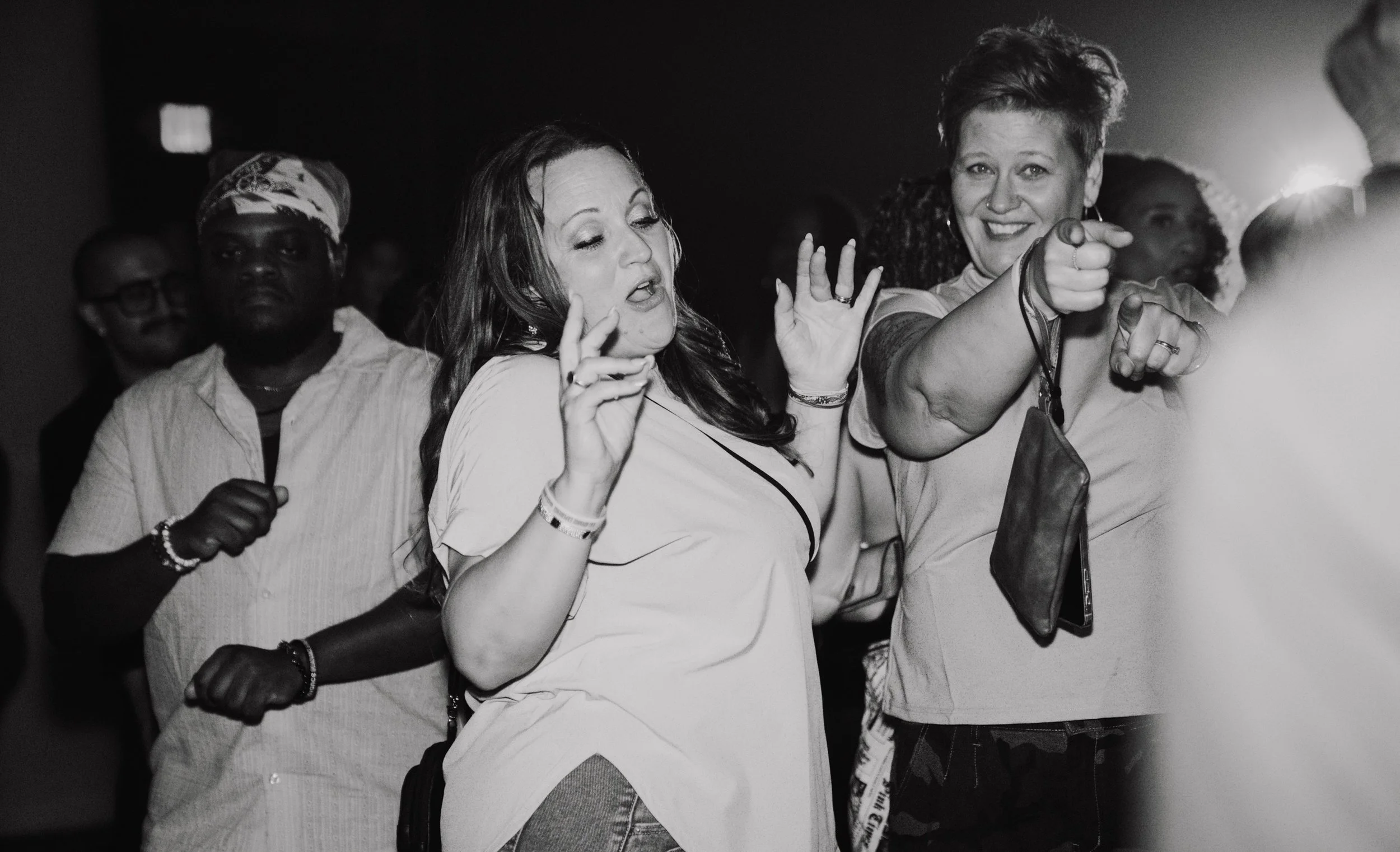 Group of women at a social event, some smiling and dancing, one woman pointing towards the camera, in a black and white photo.