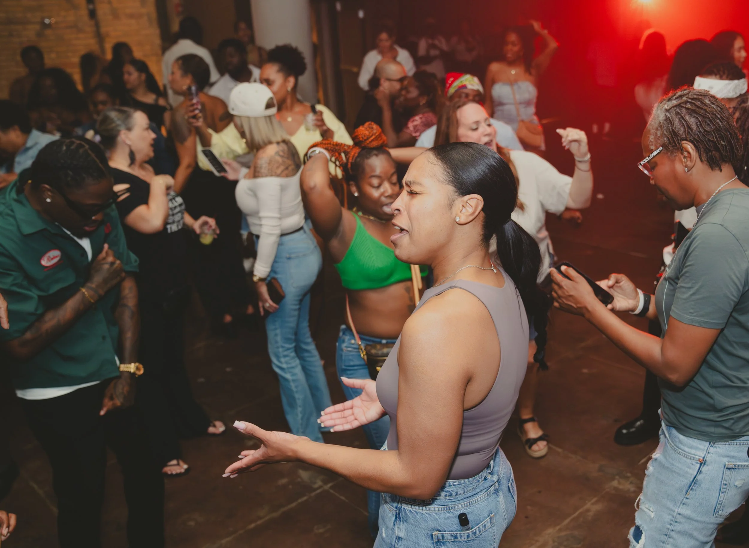 People dancing and socializing inside a dimly lit venue with red lighting, brick wall, and wooden floor.