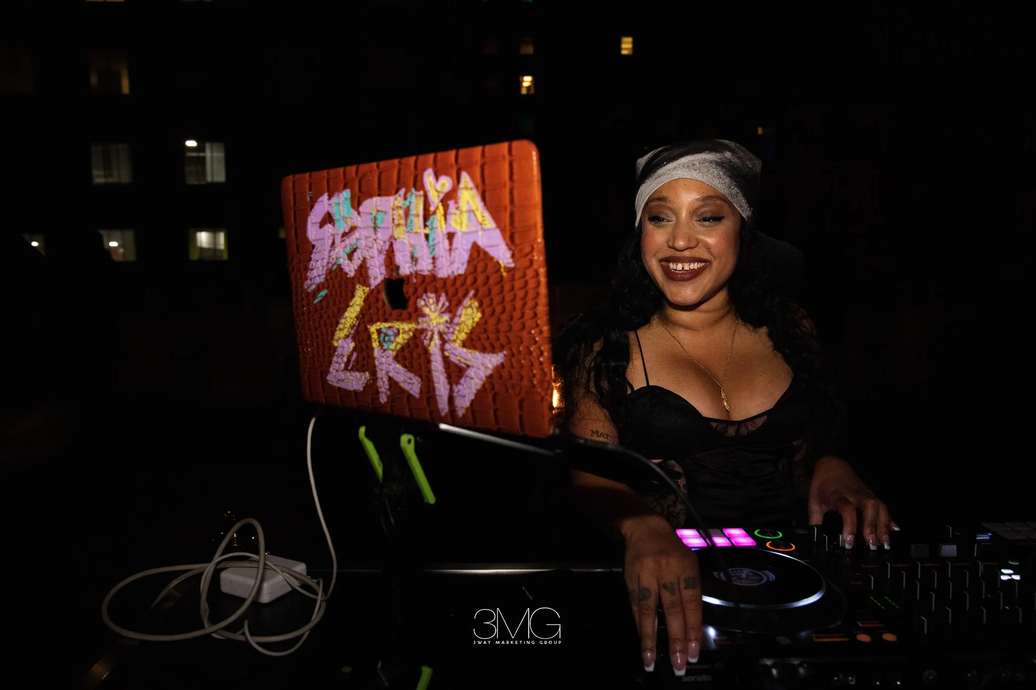 A woman with dark curly hair wearing a headscarf and black lace top is DJing at night, smiling and looking at her equipment. She is using a DJ controller with glowing buttons, and there is a red decorative sign with colorful writing partially visible