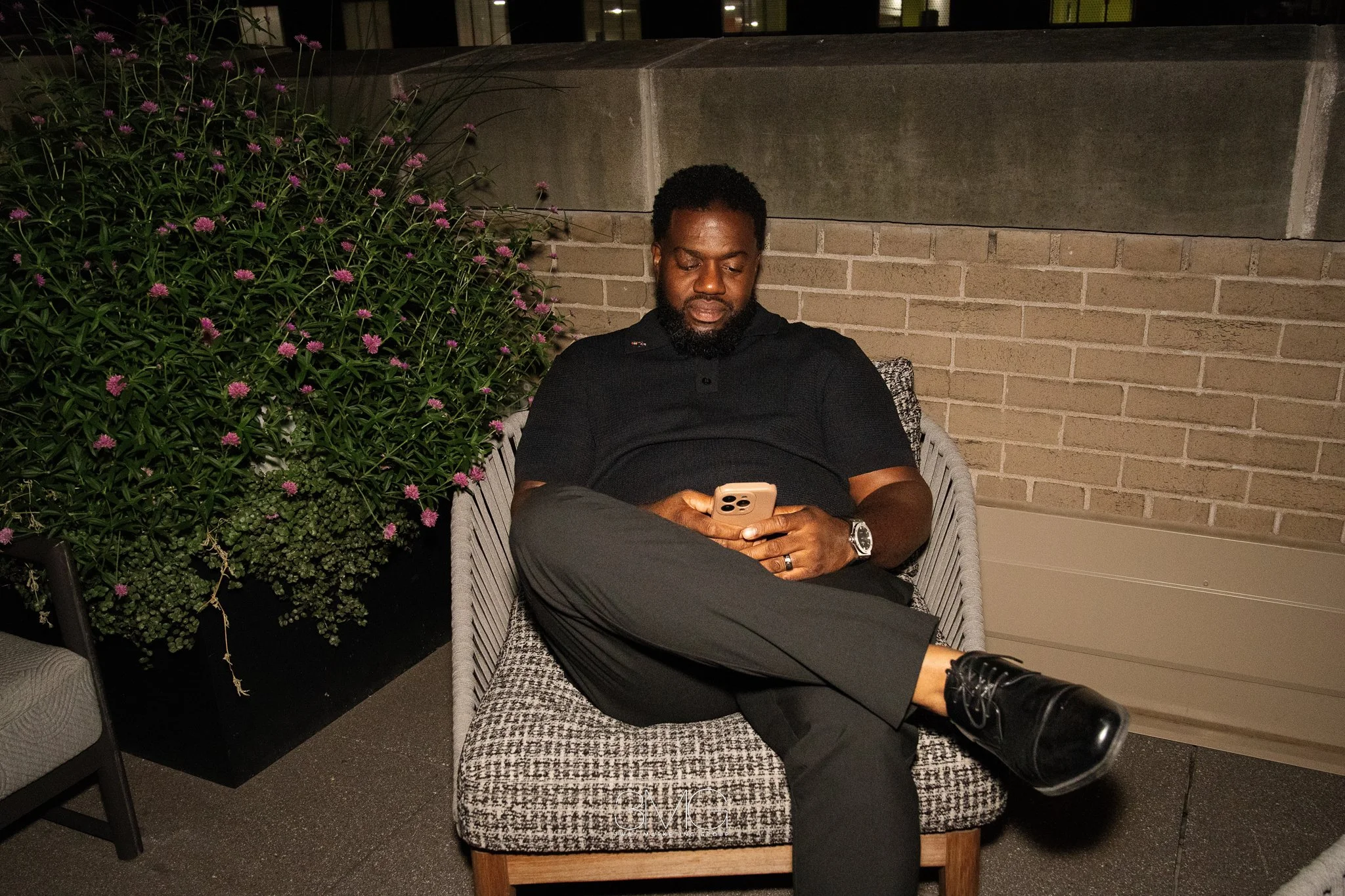 A man with a beard and curly hair sitting on a cushioned chair at night, outdoors, looking at his phone with a neutral expression. He's wearing a black polo shirt, black pants, and a wristwatch. There is a large green bush with purple flowers and a b