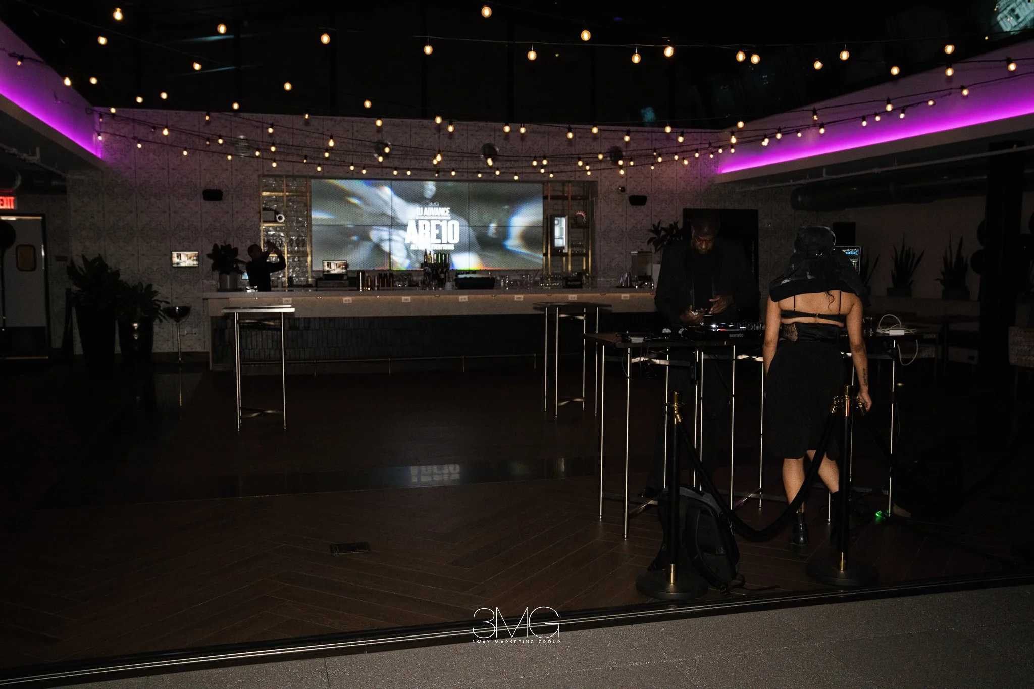 An indoor nightlife scene with a stage and DJ equipment, illuminated by string lights and purple LED accent lighting, featuring people preparing for a performance or event.