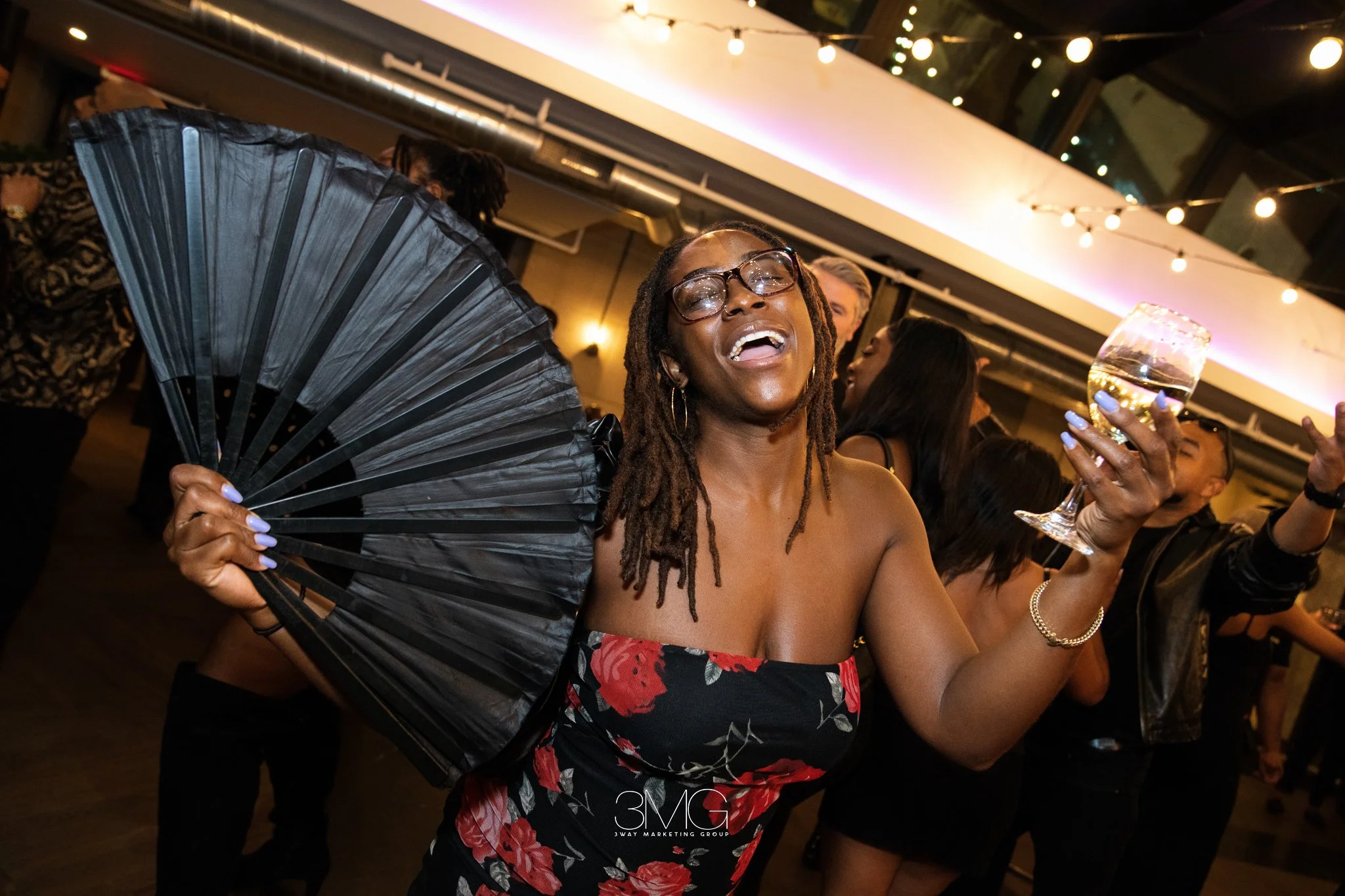 A woman with glasses and dreadlocks smiling and enjoying a celebration at night, holding a glass of white wine, surrounded by other people, with string lights overhead.