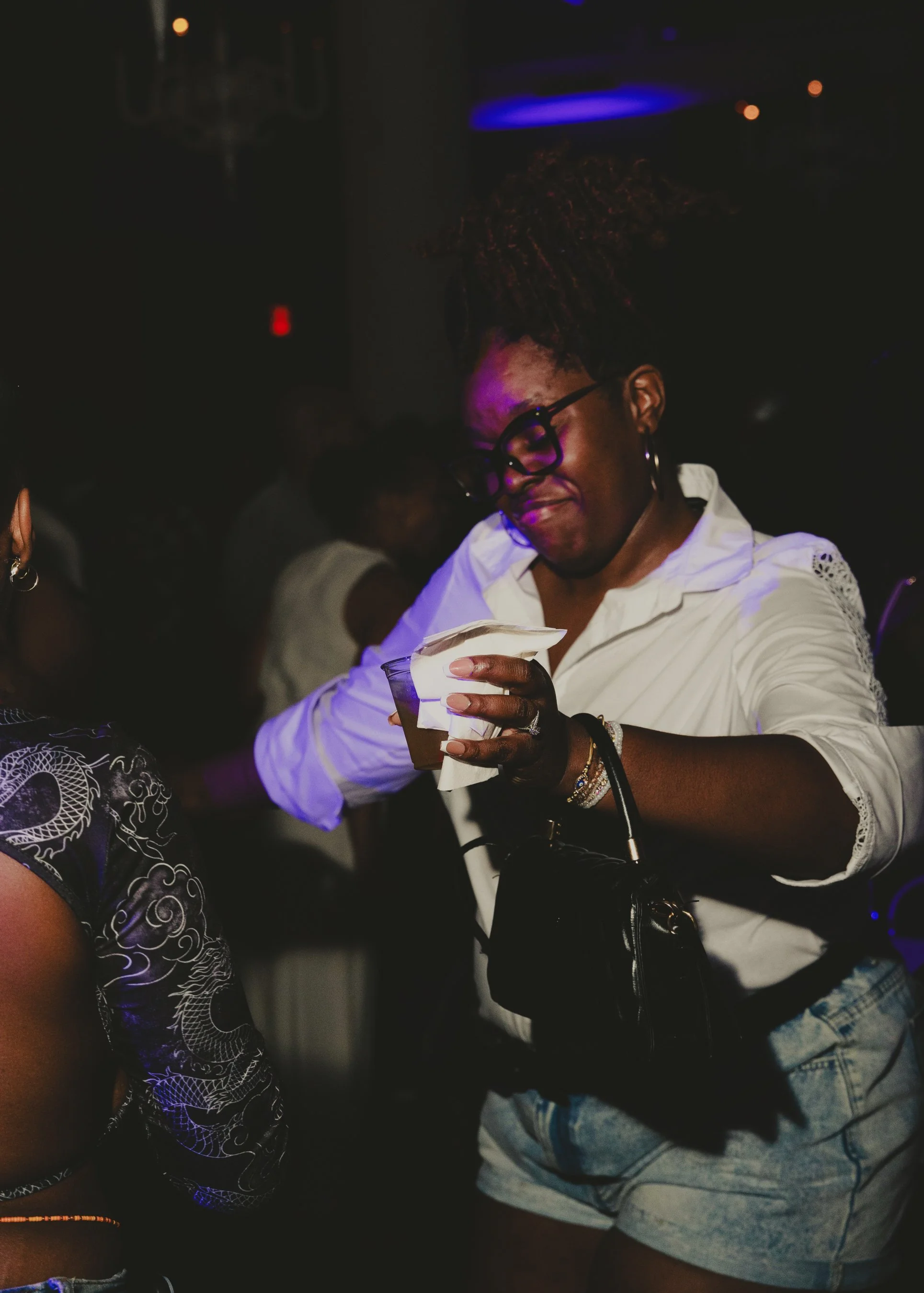 A woman with glasses and hoop earrings in a white shirt, holding a drink and a napkin, in a dark indoor setting with colorful lighting.
