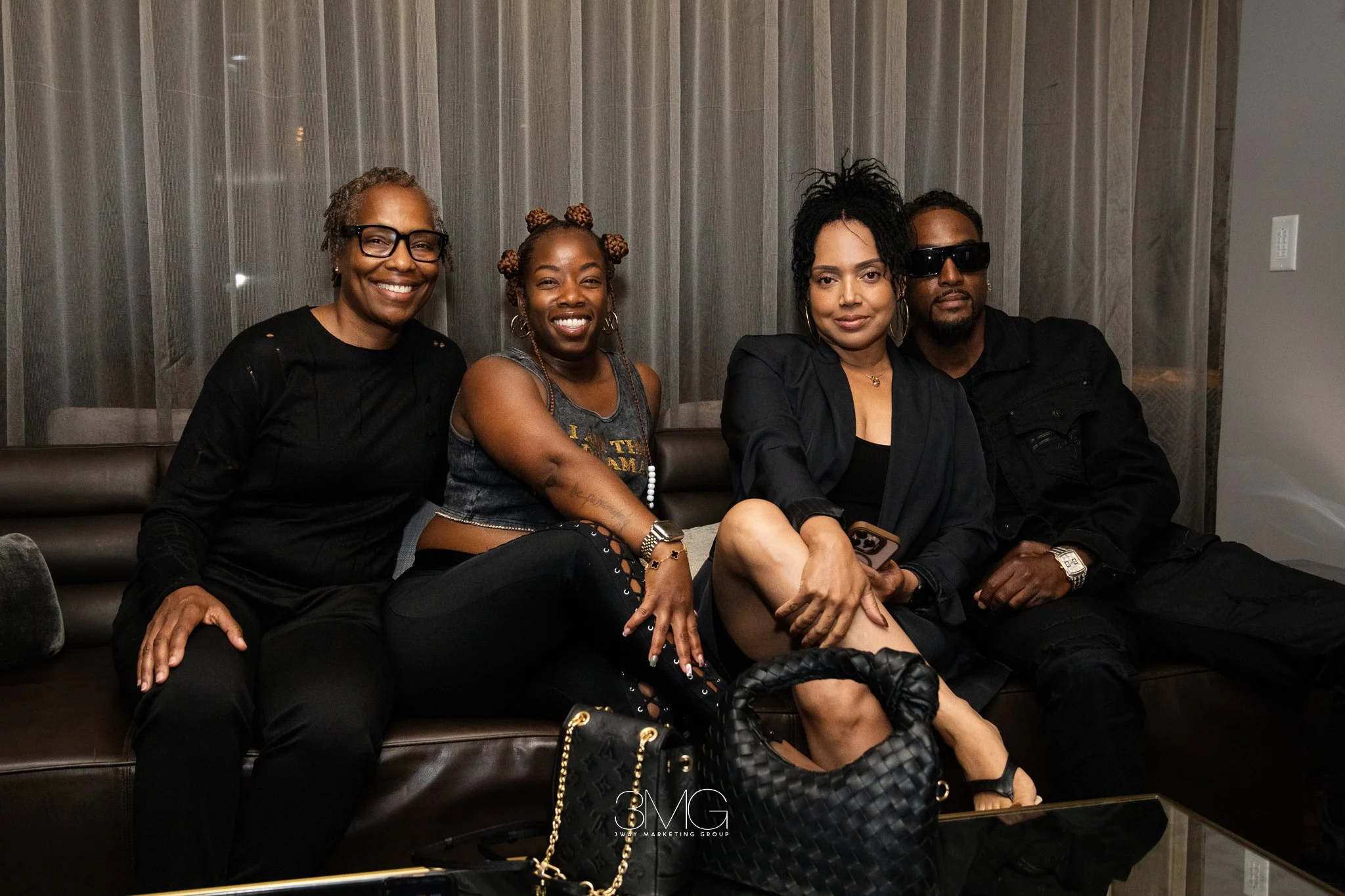 Four people sitting on a brown leather couch in front of a sheer curtain, smiling and posing for the photo. Two women and two men, with one woman wearing glasses, another with bun hairstyles, and a man with sunglasses.