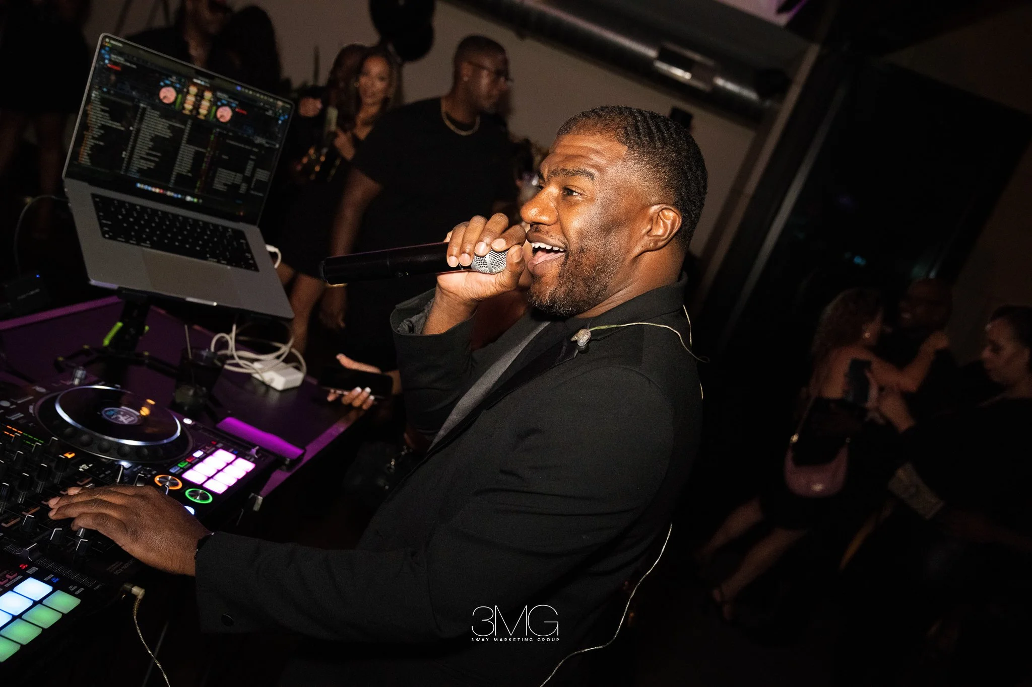 A man with short hair and a beard enjoying himself as he uses a DJ controller at a party, holding a microphone near his mouth, with people dancing in the background.
