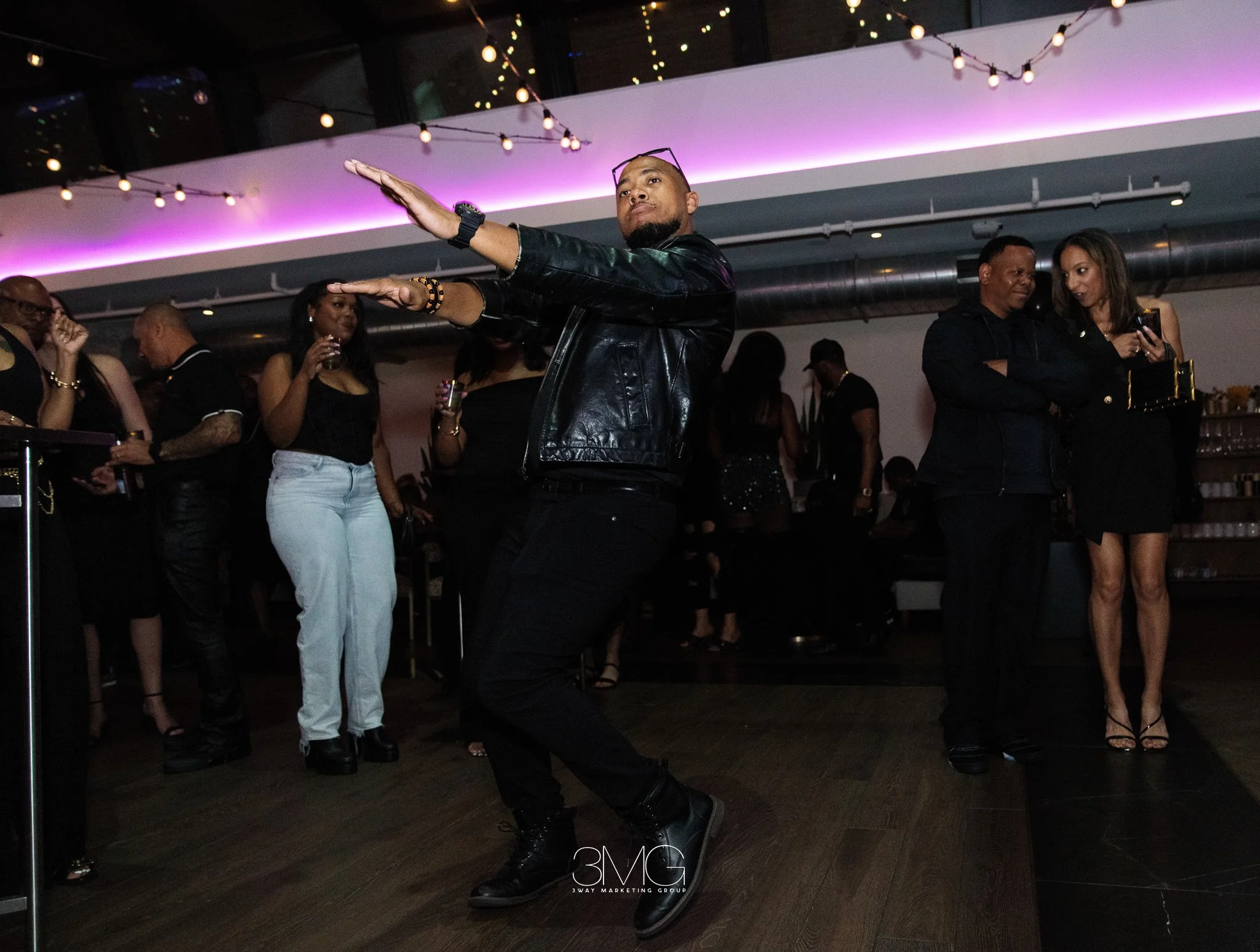 A man dancing at a social gathering or party, surrounded by people, with colorful lighting and string lights overhead.