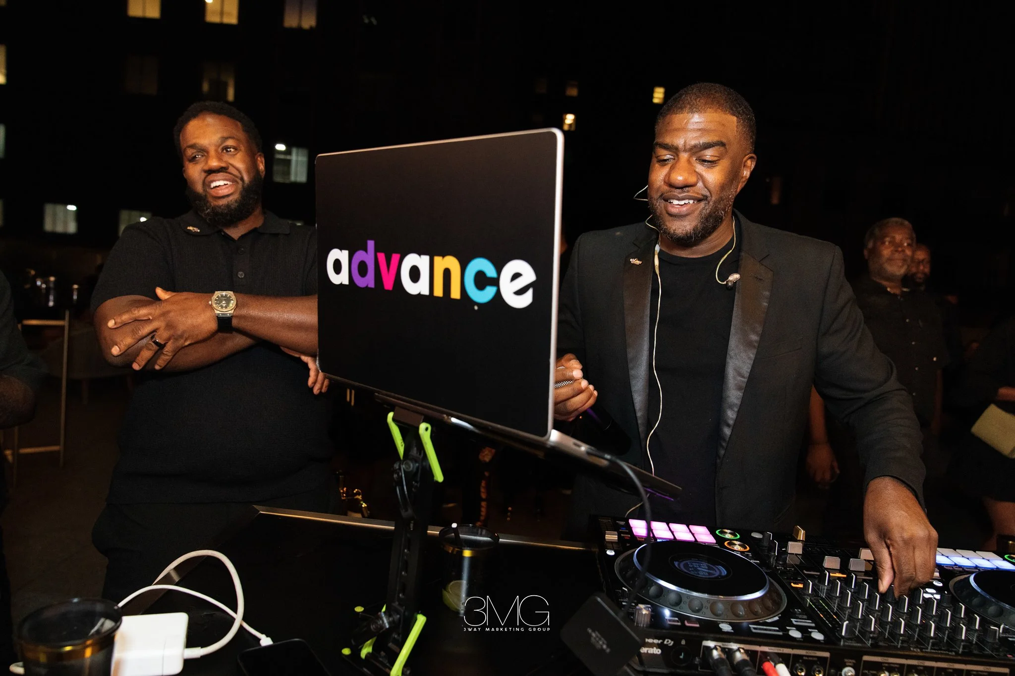 Two men at a nighttime outdoor event with DJ equipment. One man is wearing a black t-shirt, has a beard, and is smiling and looking at the DJ equipment. The other man, also with a beard, is wearing a black blazer and is focused on the DJ gear. The DJ