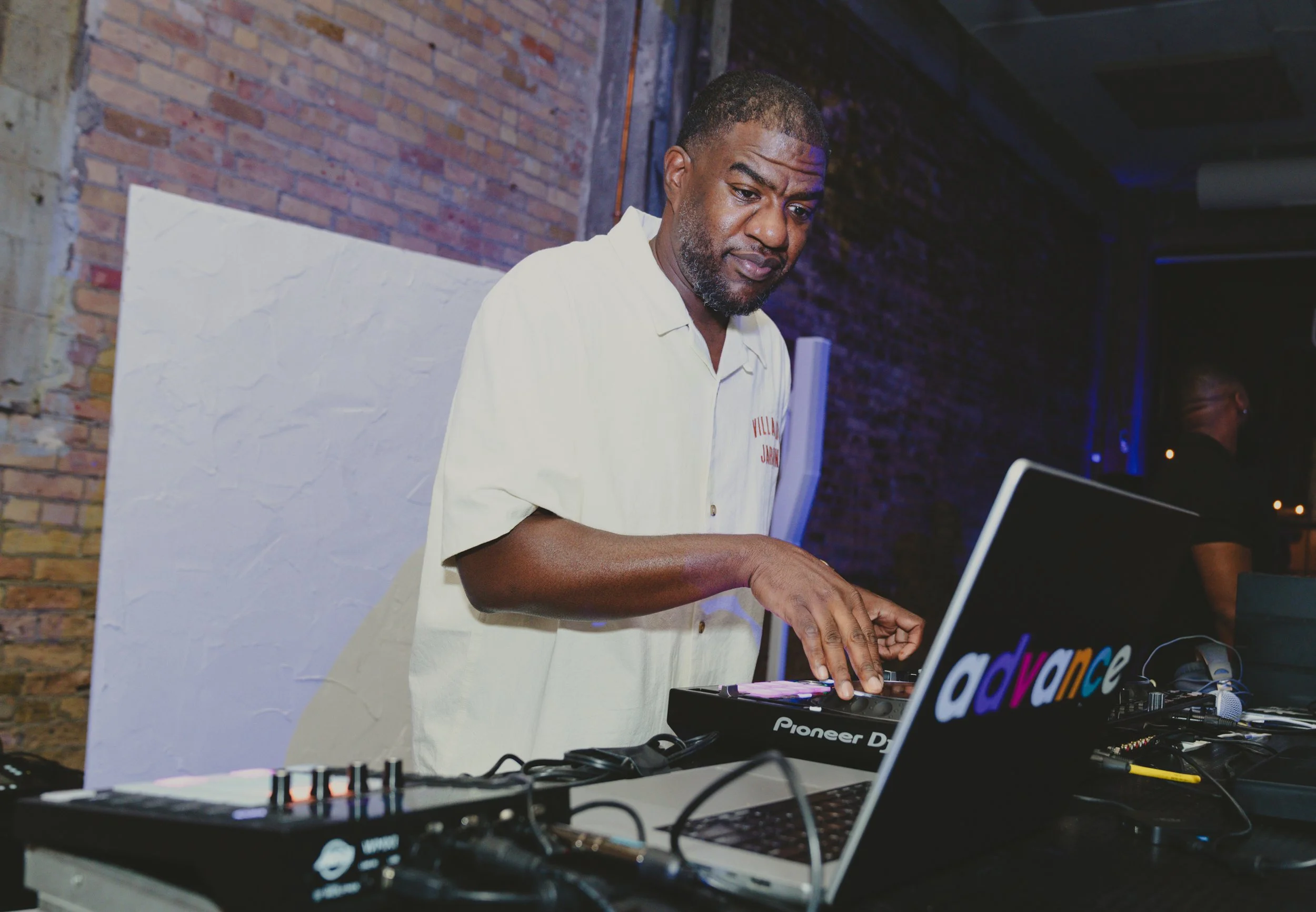 A man with short dark hair and a beard wearing a white shirt, standing at a DJ setup, in a room with brick walls, with a focused expression.