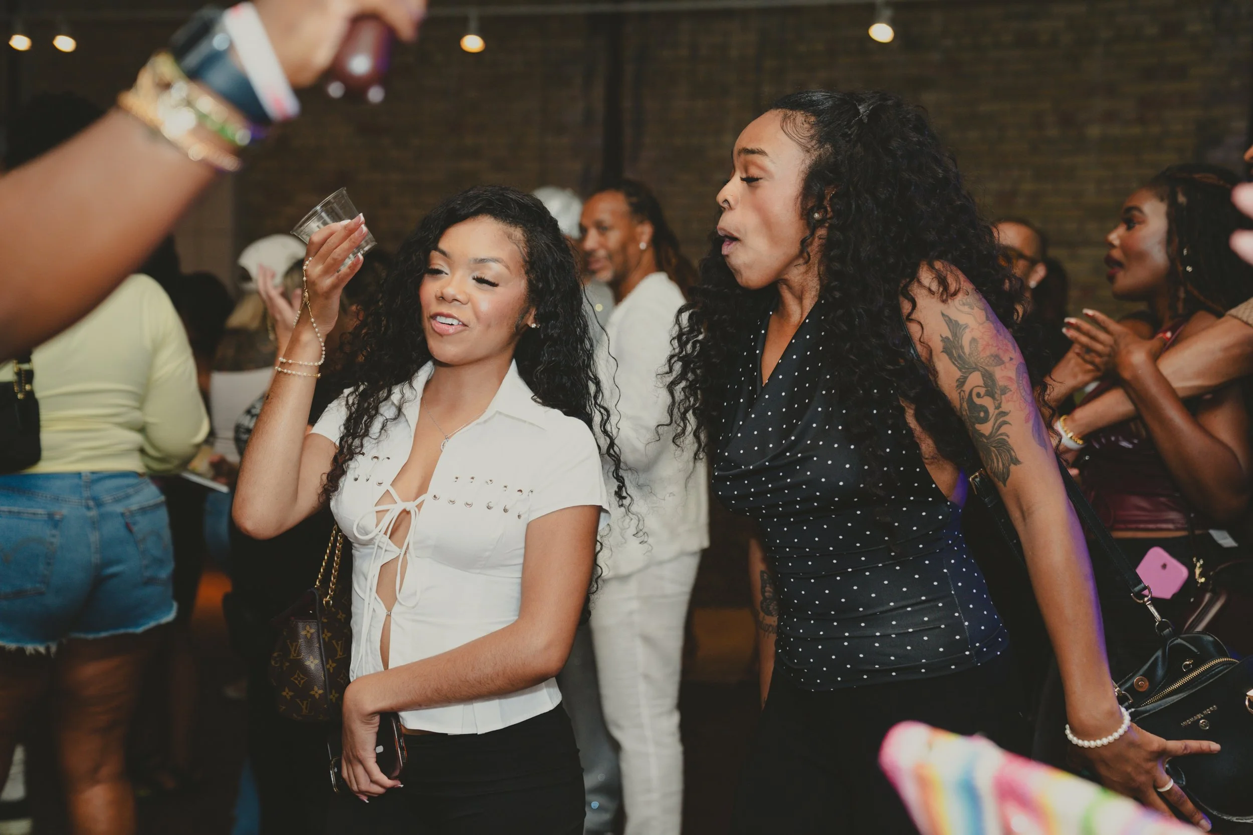 Group of women socializing at an indoor gathering, with some holding drinks, in front of a brick wall.