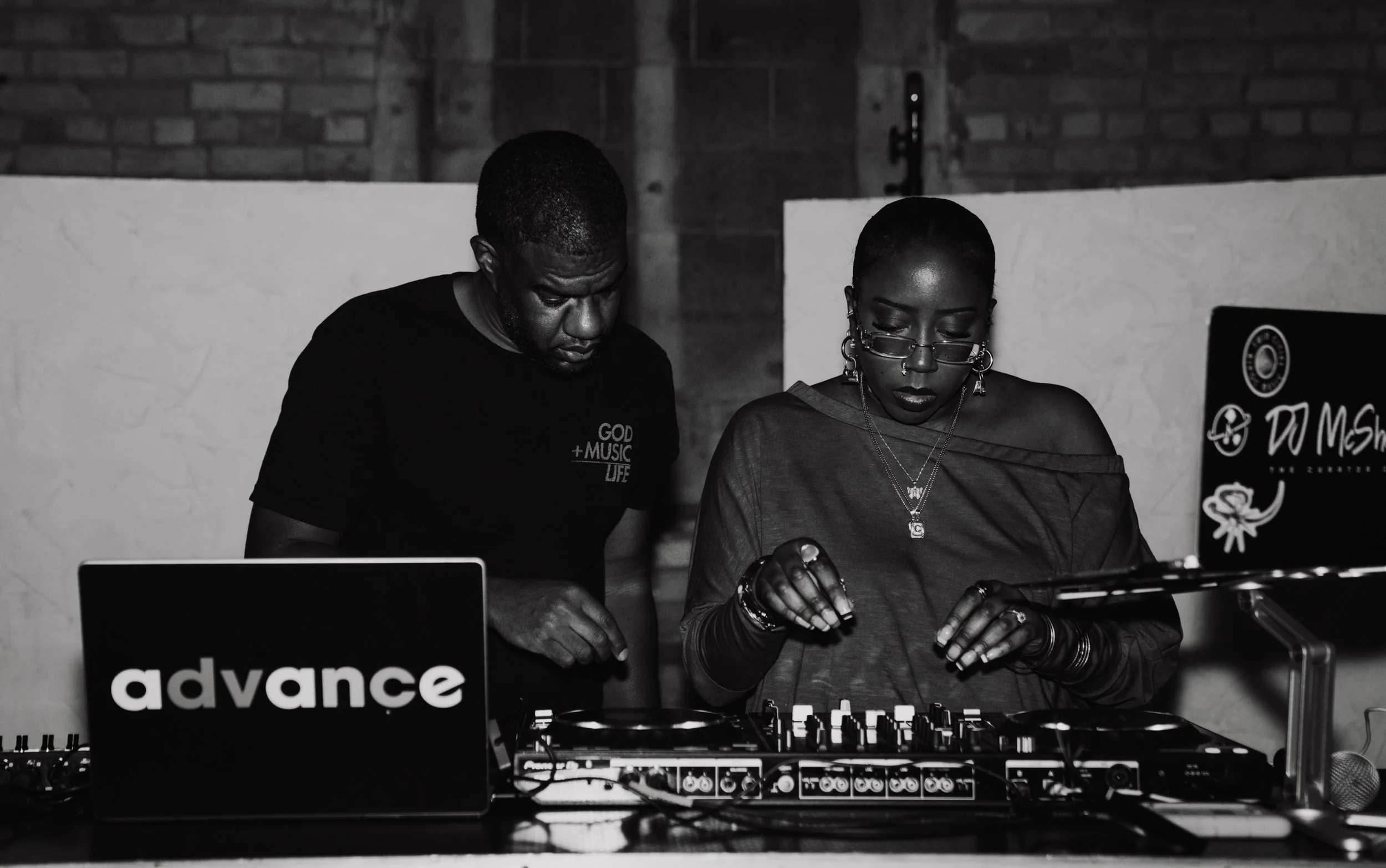 Two people working at a DJ setup with a laptop and turntables, in a dimly lit room with a brick wall background. One person is wearing glasses and has short hair, the other has short curly hair. There's a sign that says "advance" in the foreground.