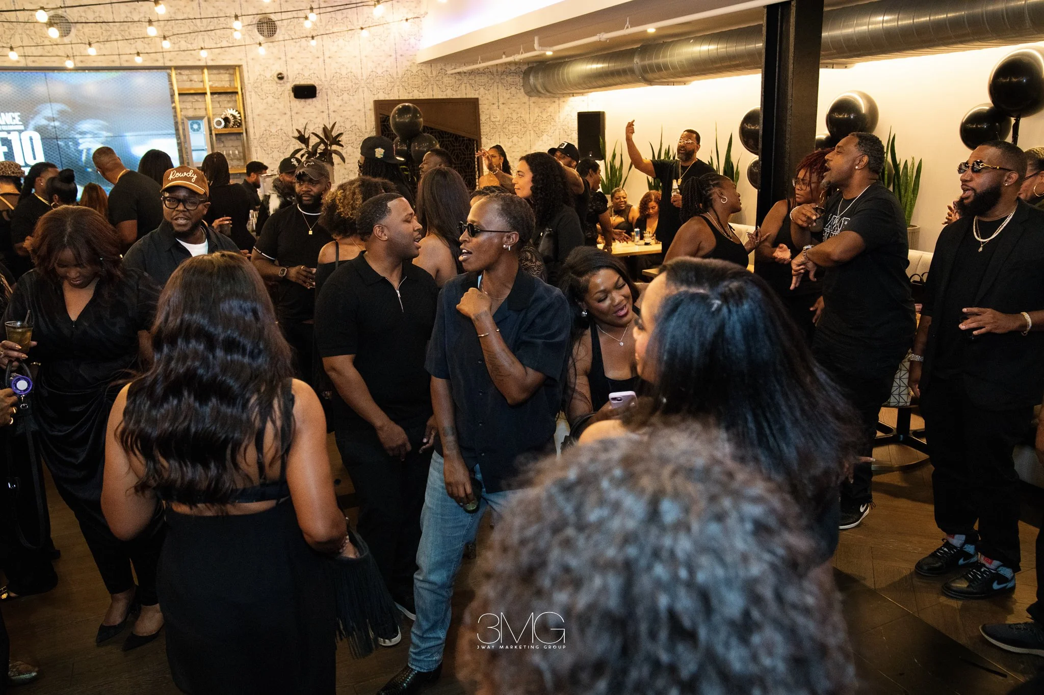 A crowded indoor party with people chatting, dancing, and enjoying themselves, with a large screen on the wall displaying the word 'F10' and decorative balloons around the room.
