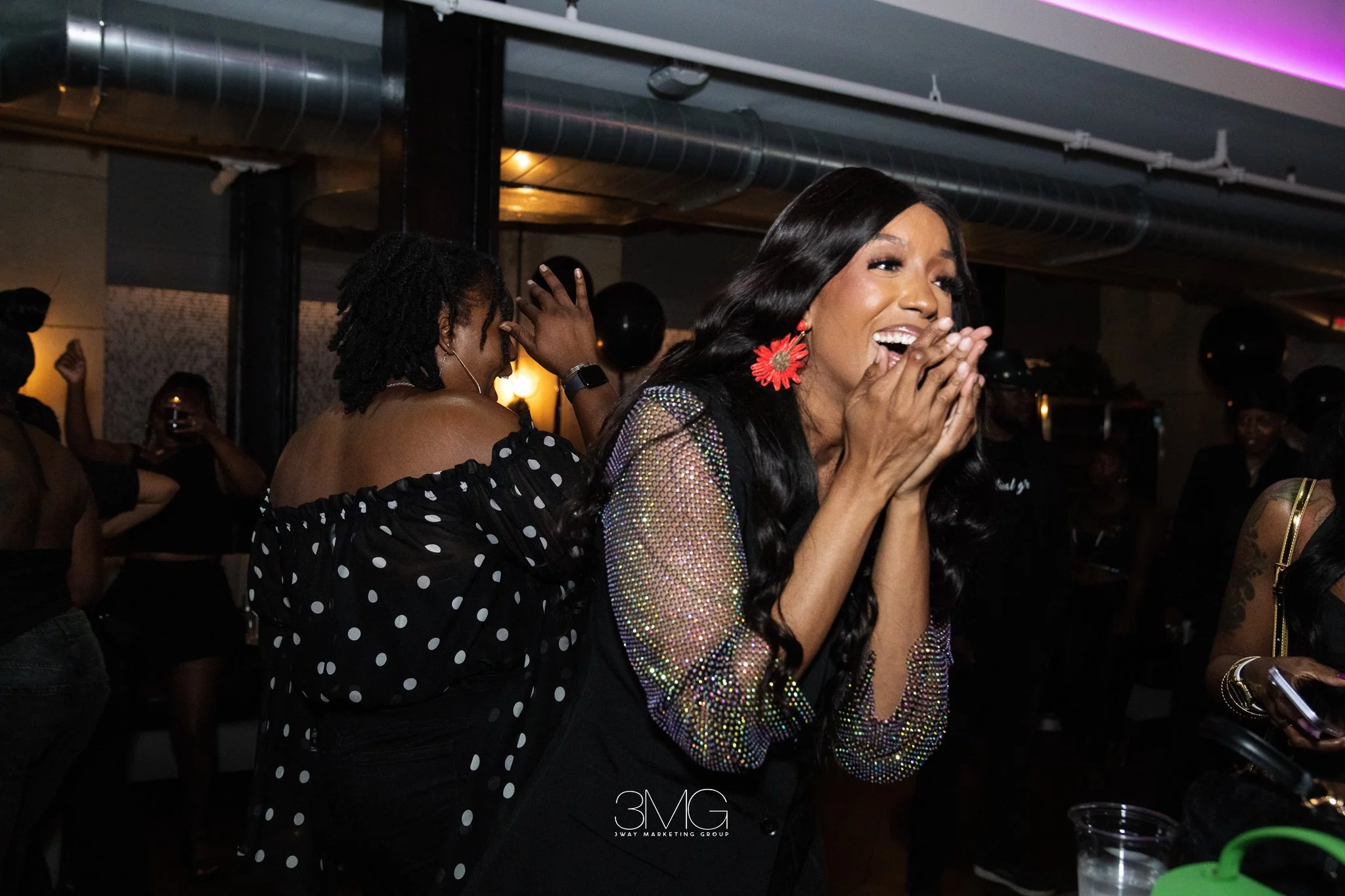 A woman with long dark hair, wearing red flower earrings and a shimmering top, appears surprised or excited at a party or celebration, surrounded by people.