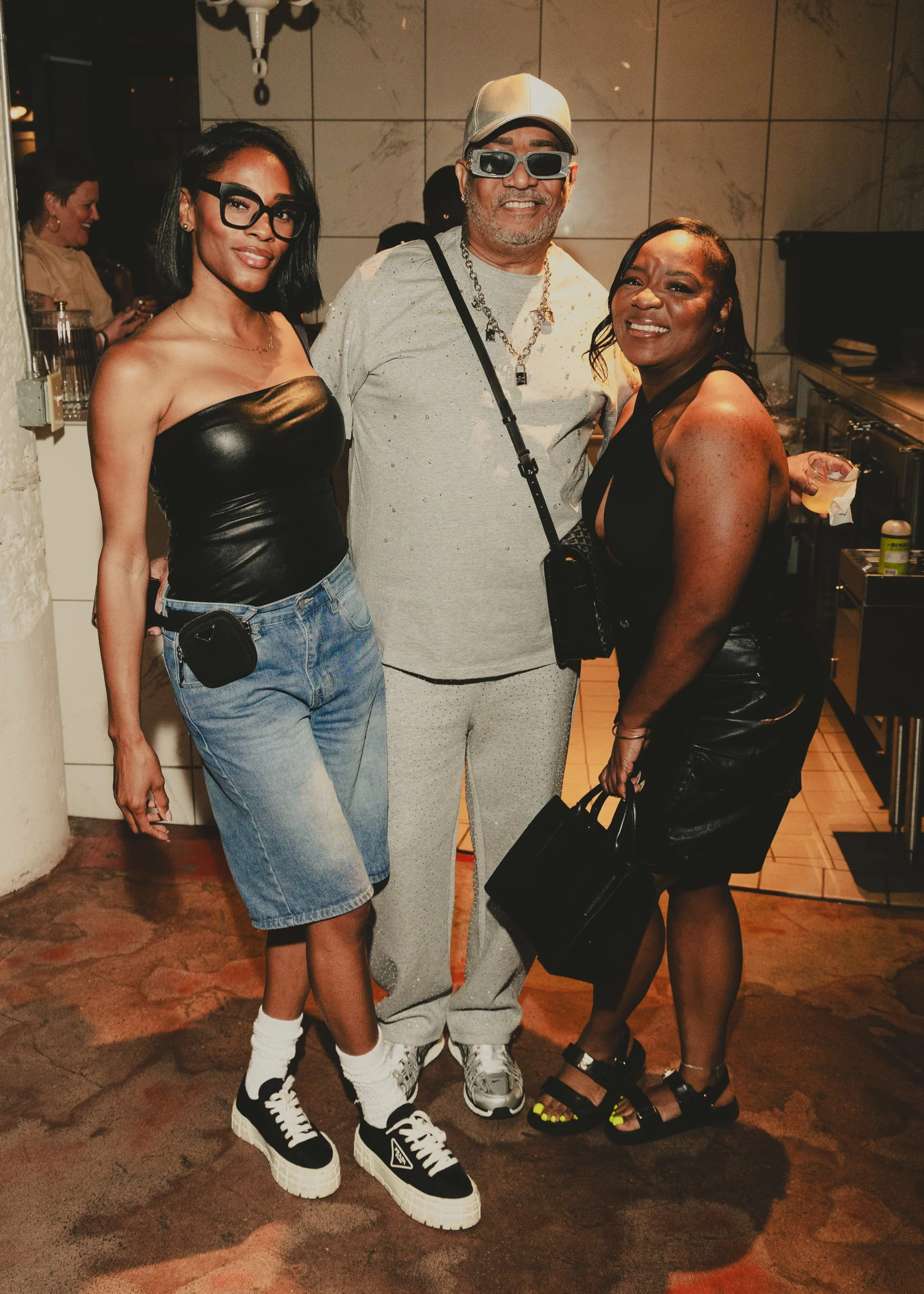Three people posing together indoors, two women and one man. The woman on the left has shoulder-length black hair, glasses, a black leather top, denim shorts, and sneakers. The man in the middle has a beard, sunglasses, a bucket hat, a chain necklace
