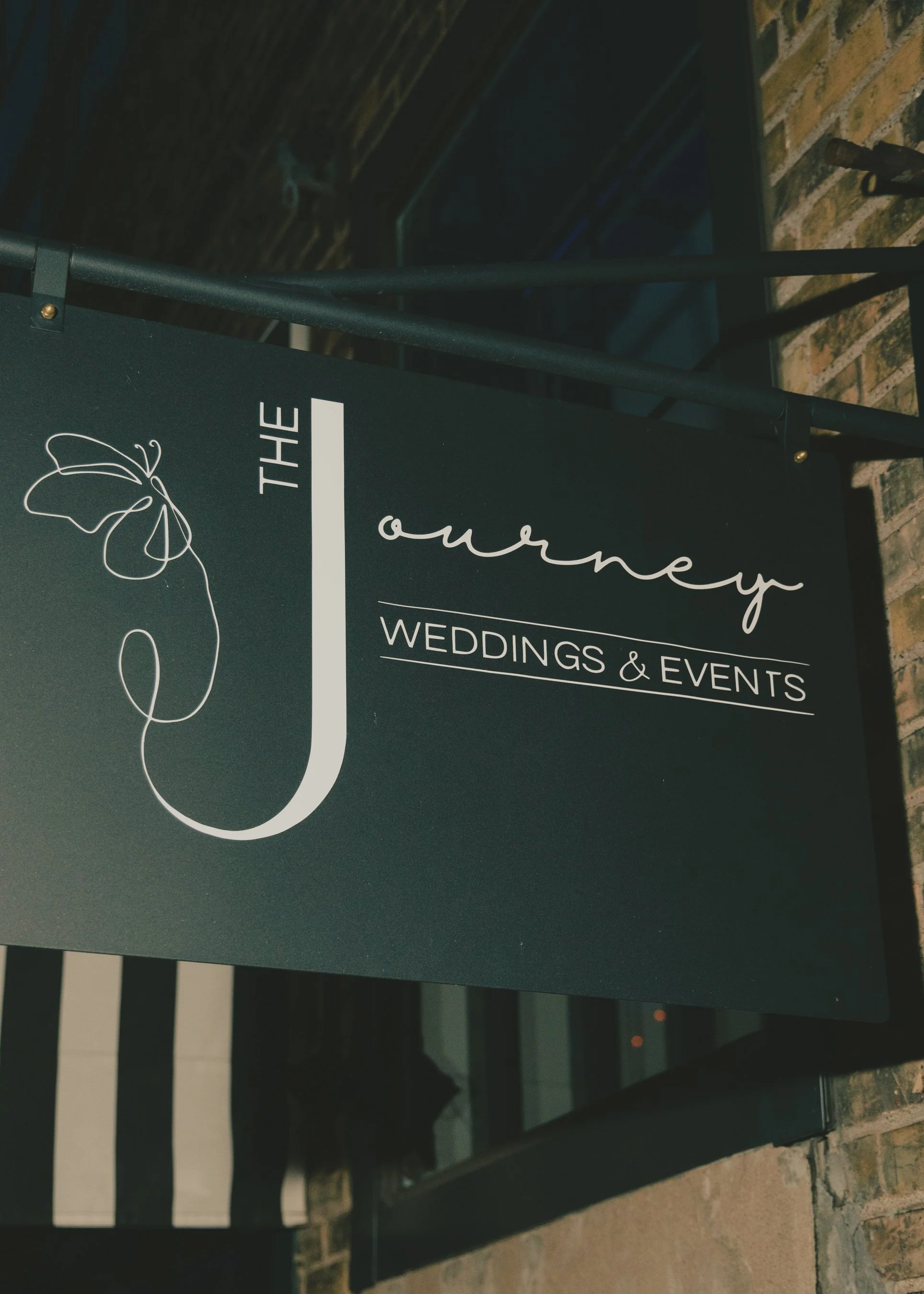 A black sign with white cursive text reading 'The Oreny Weddings & Events' and a line drawing of a butterfly, attached to a black metal railing outside against a brick wall.