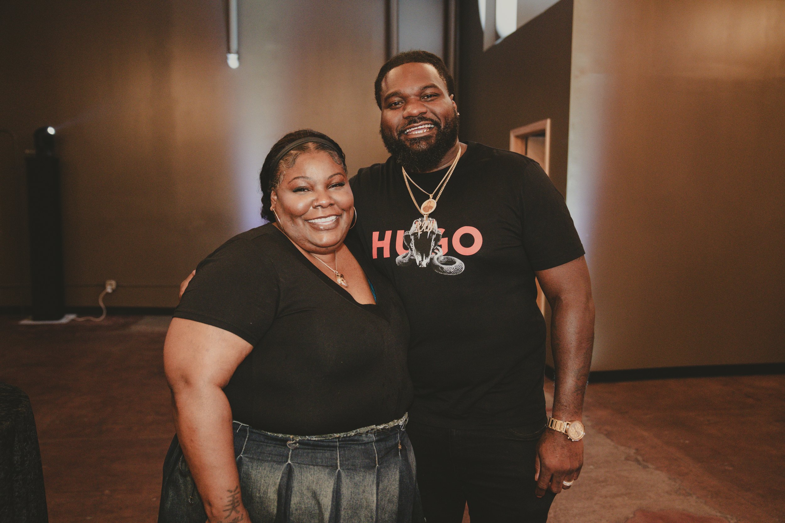 A smiling woman and man standing close together in an indoor setting. The woman wears a black top and jeans, and the man wears a black t-shirt with a graphic design and layered gold necklaces. They appear happy and are posing for the photo.