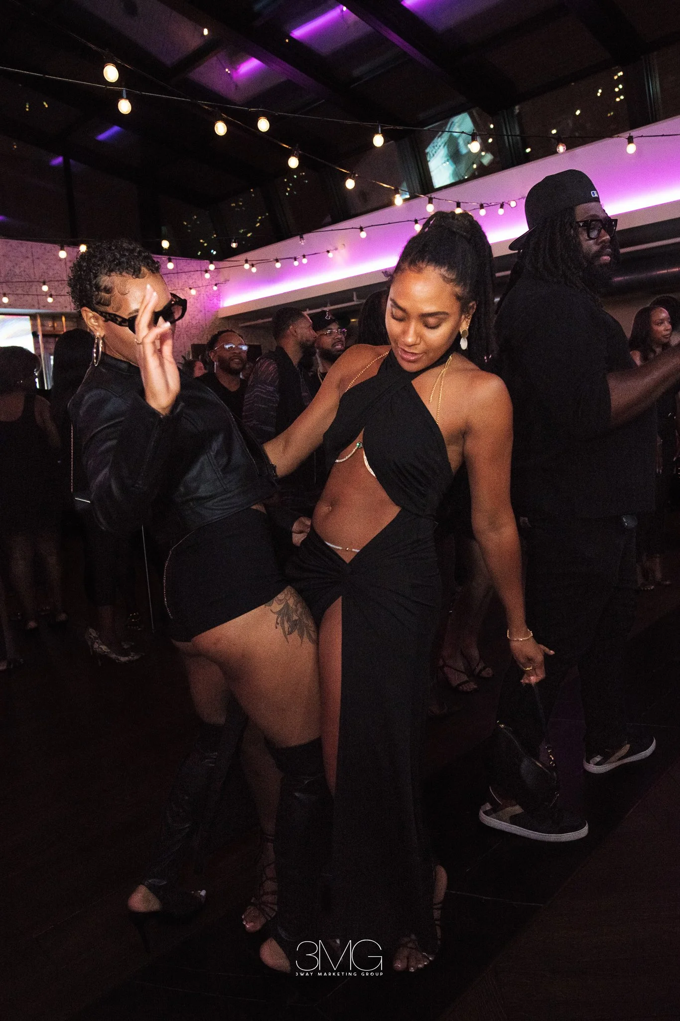 Two women dancing at a club with purple neon lights and string lights. One woman is wearing a black dress with a side slit, and the other is wearing a black jacket and shorts. The background has other people dancing and enjoying the nightclub atmosph