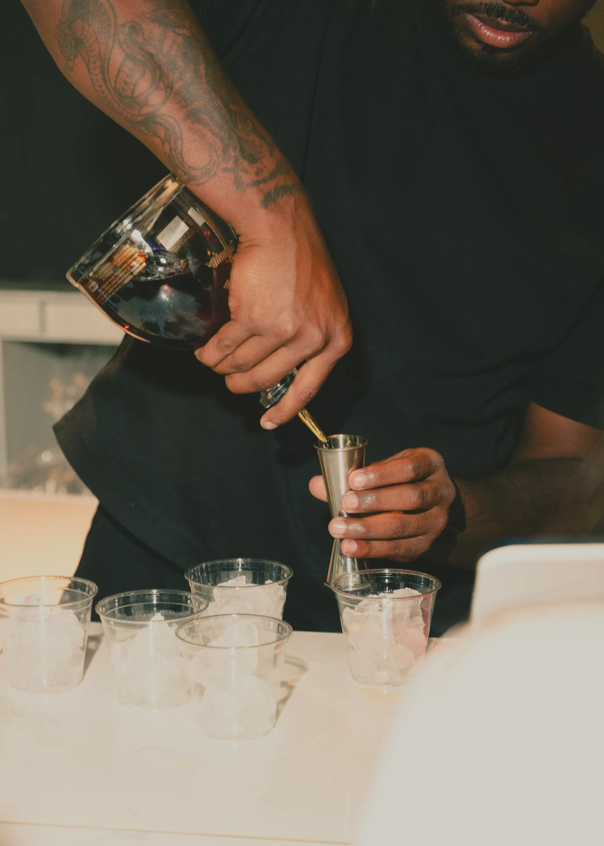 A person pouring a drink from a bottle into a small cup, with multiple empty cups on the table in front of them.