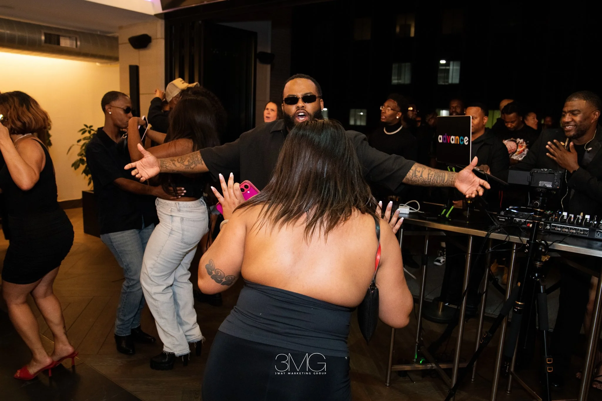 A DJ in sunglasses performing at a lively party with people dancing and socializing indoors.