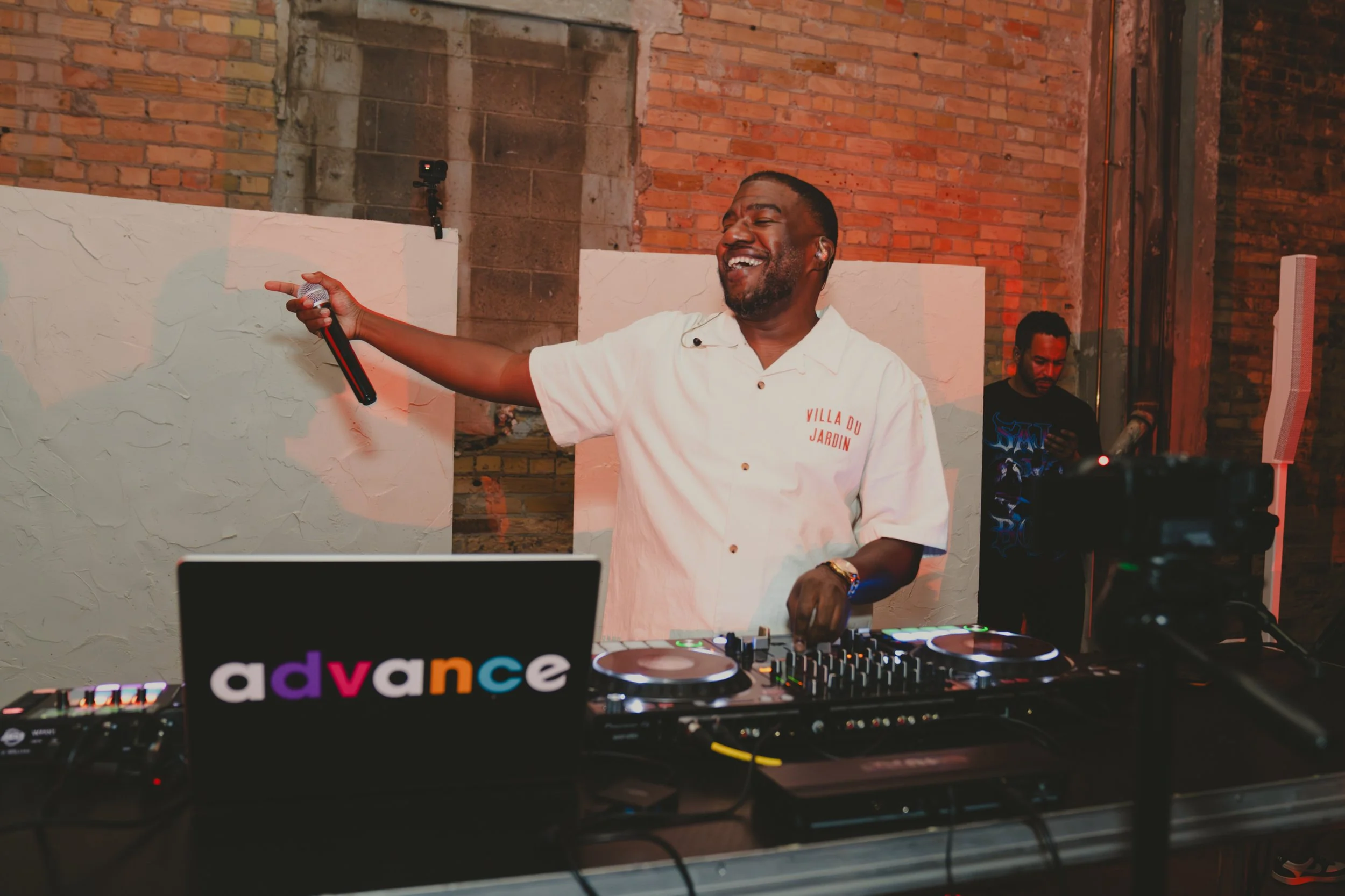 A man with short black hair, a beard, and a wide smile is DJing at an event. He is wearing a white shirt with red text on it and holding a microphone in his right hand. He is standing behind a DJ setup with a laptop displaying a colorful 'advance' lo