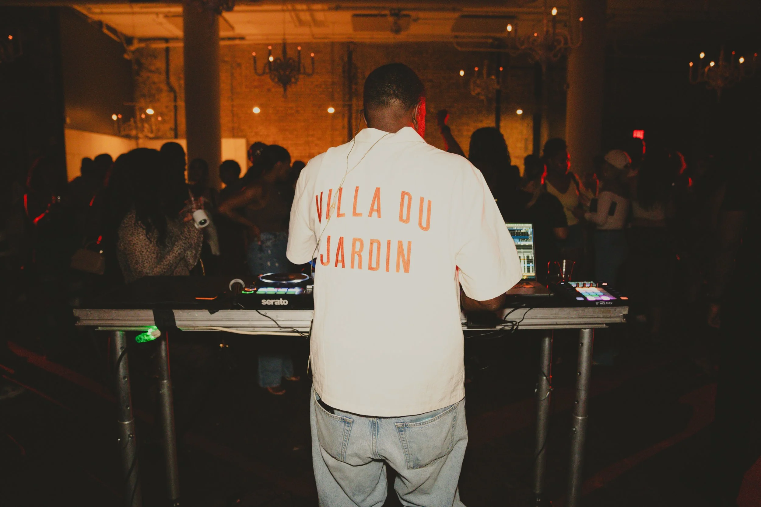 A DJ wearing a white shirt with orange text that reads 'VILLA DU JARDIN' supervises music equipment in a dimly lit nightclub or party setting with brick walls, chandeliers, and a crowd of people dancing.